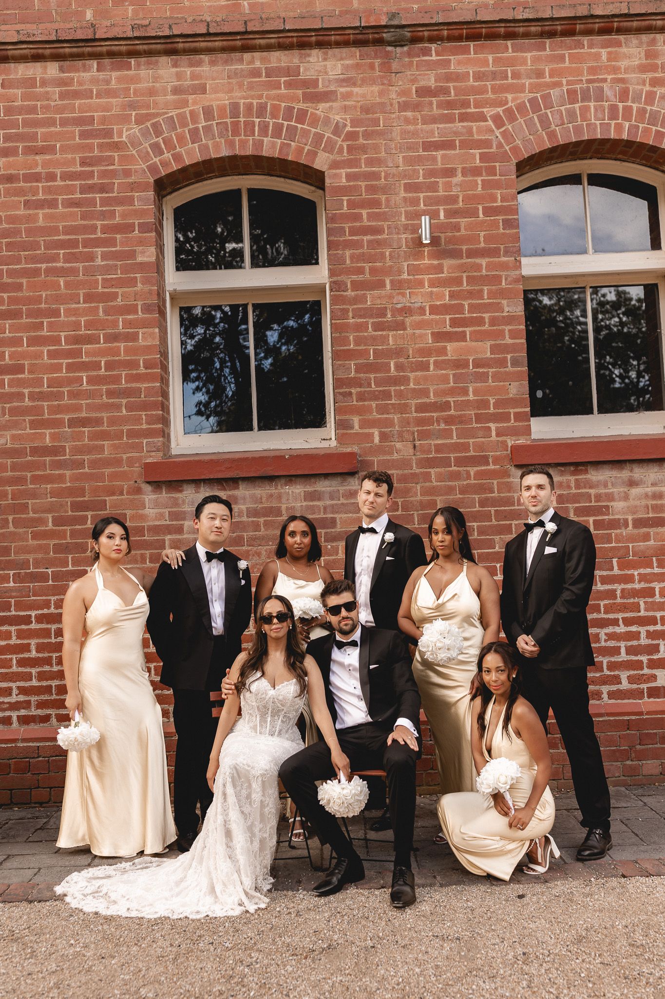 Diverse wedding party poses confidently against a red brick wall in elegant attire.