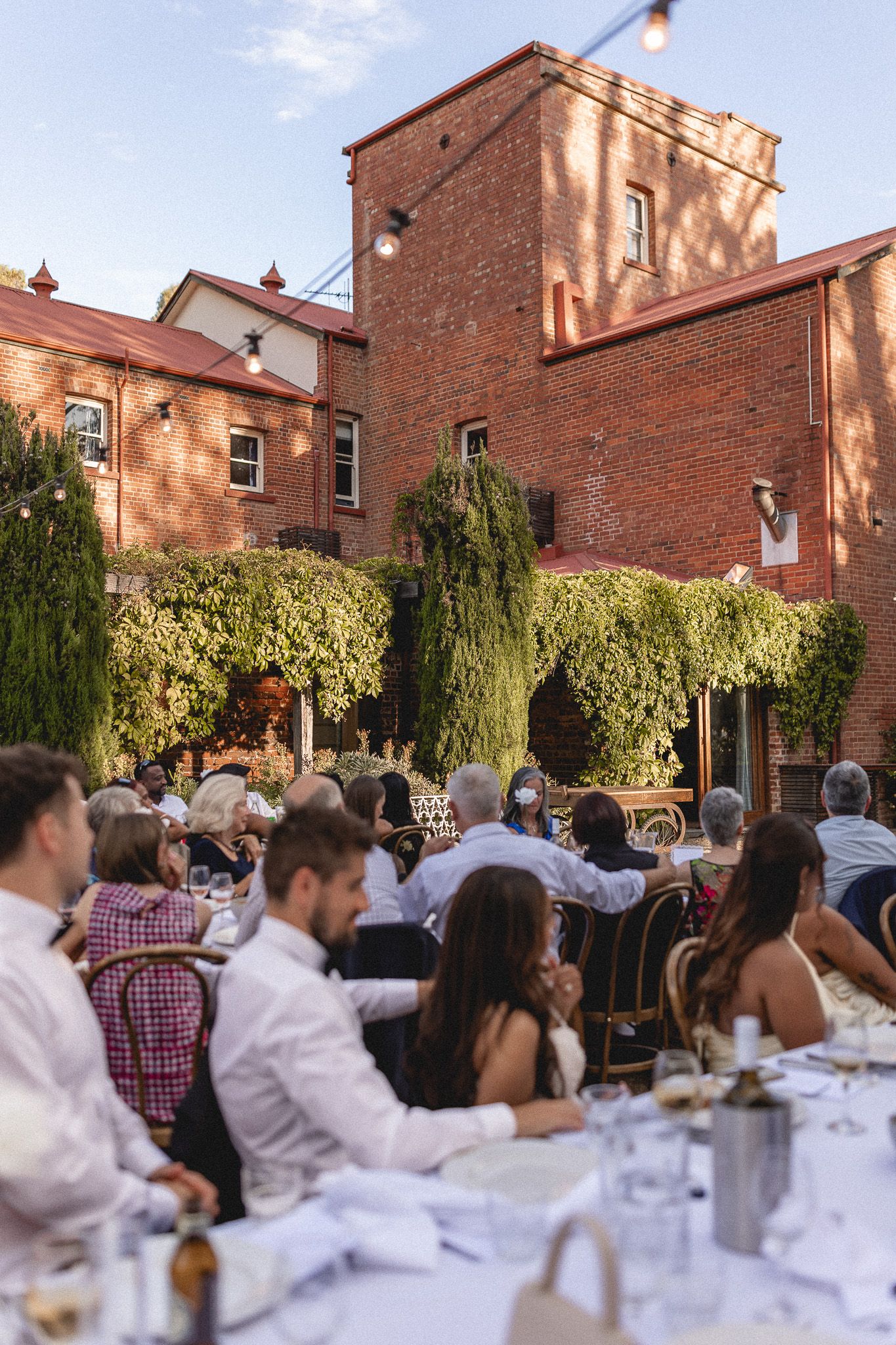 Guests dine outdoors at a wedding reception beside an ivy-covered historic brick building.