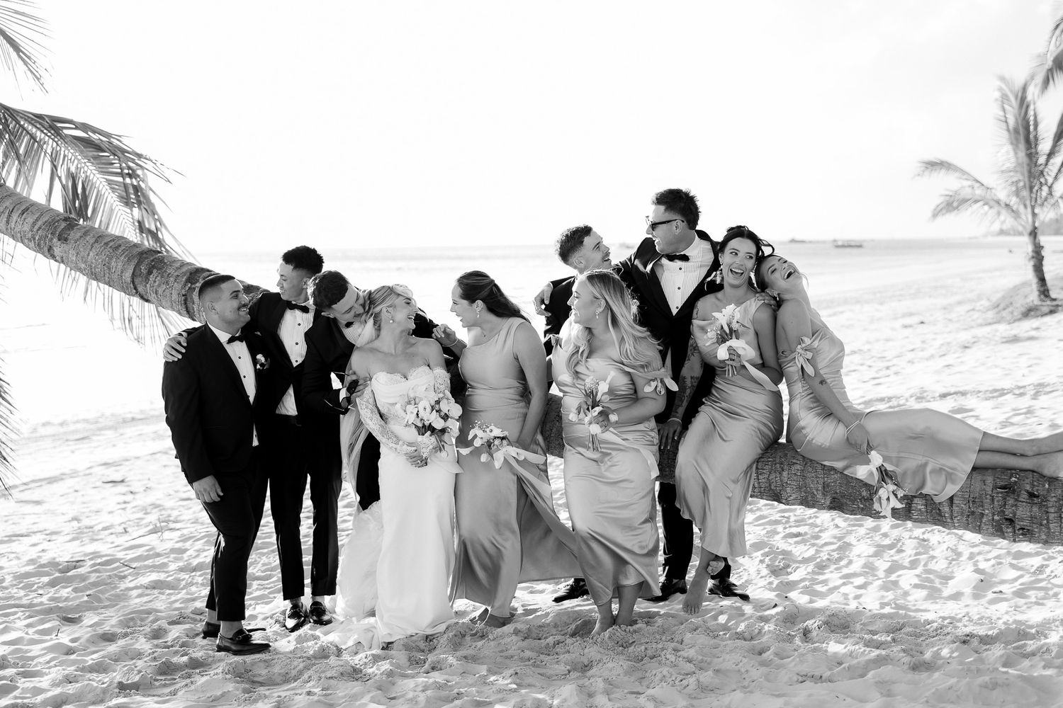 Black and white beach wedding party photo with bride, groom, and bridal party laughing on sandy shore near palm trees.