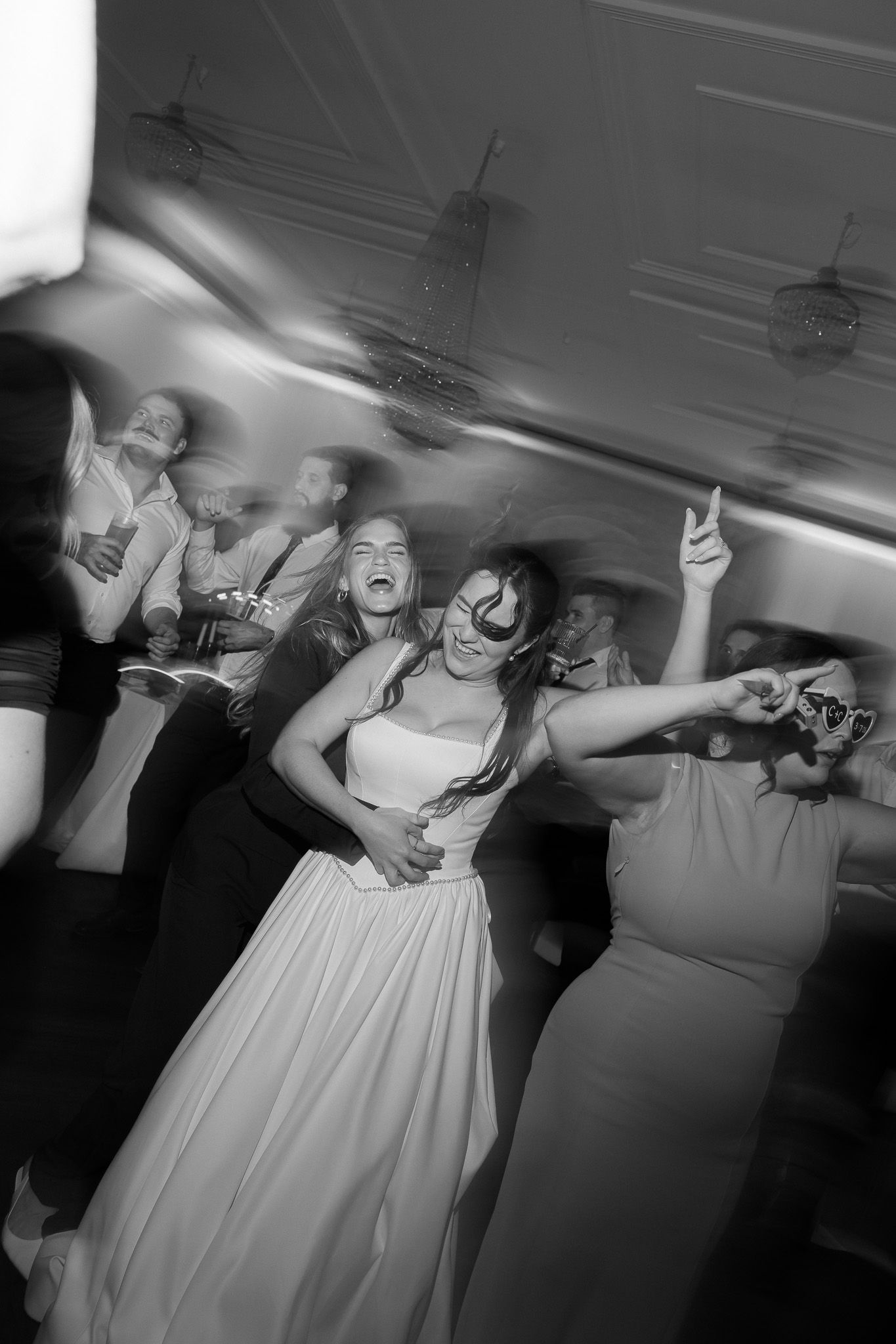 Black and white photo of a bride laughing and dancing joyfully with guests at a lively wedding reception.