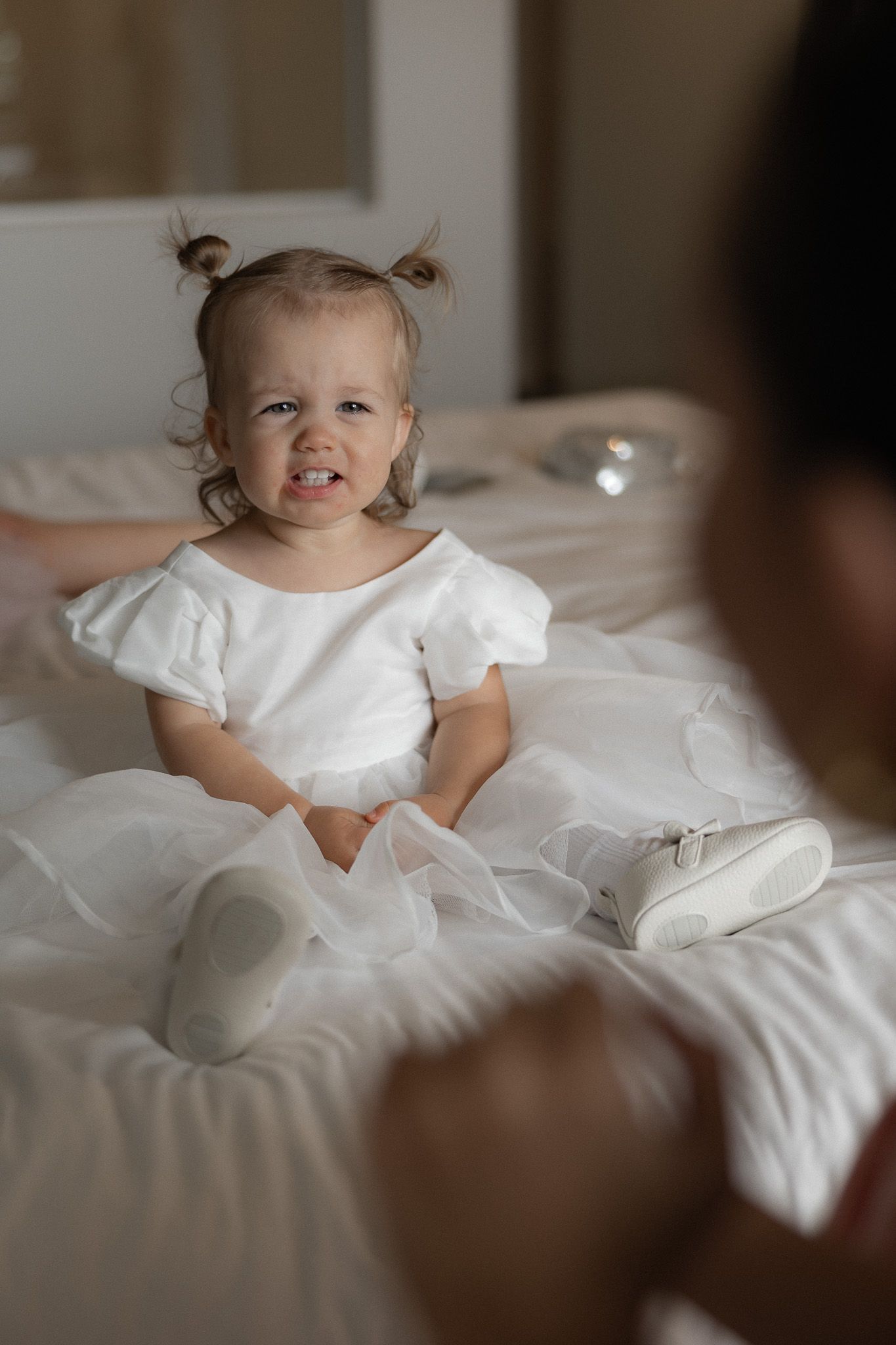 Adorable toddler flower girl in white dress sits on bed with pigtails at wedding preparation.