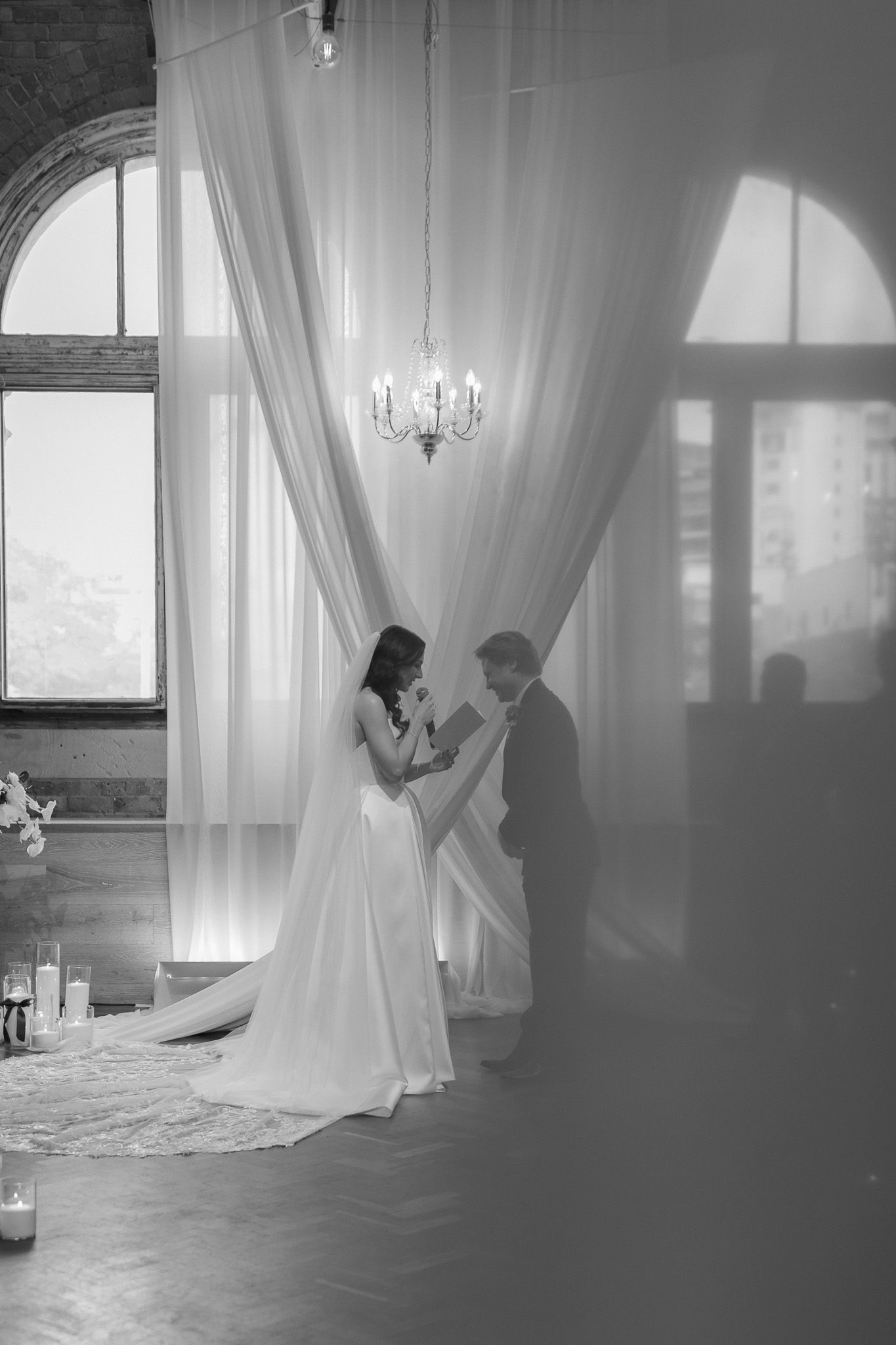 Black and white photo of bride and groom sharing an intimate moment under draped white fabric and chandelier.