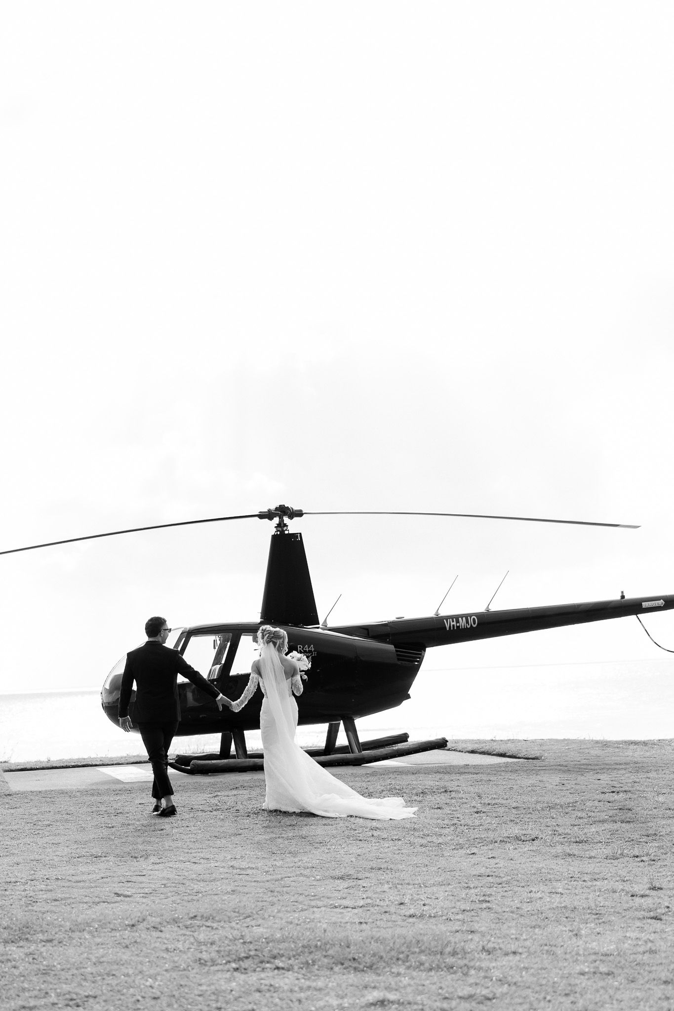 Black and white photo of a bride and groom walking hand in hand toward a helicopter on their wedding day.