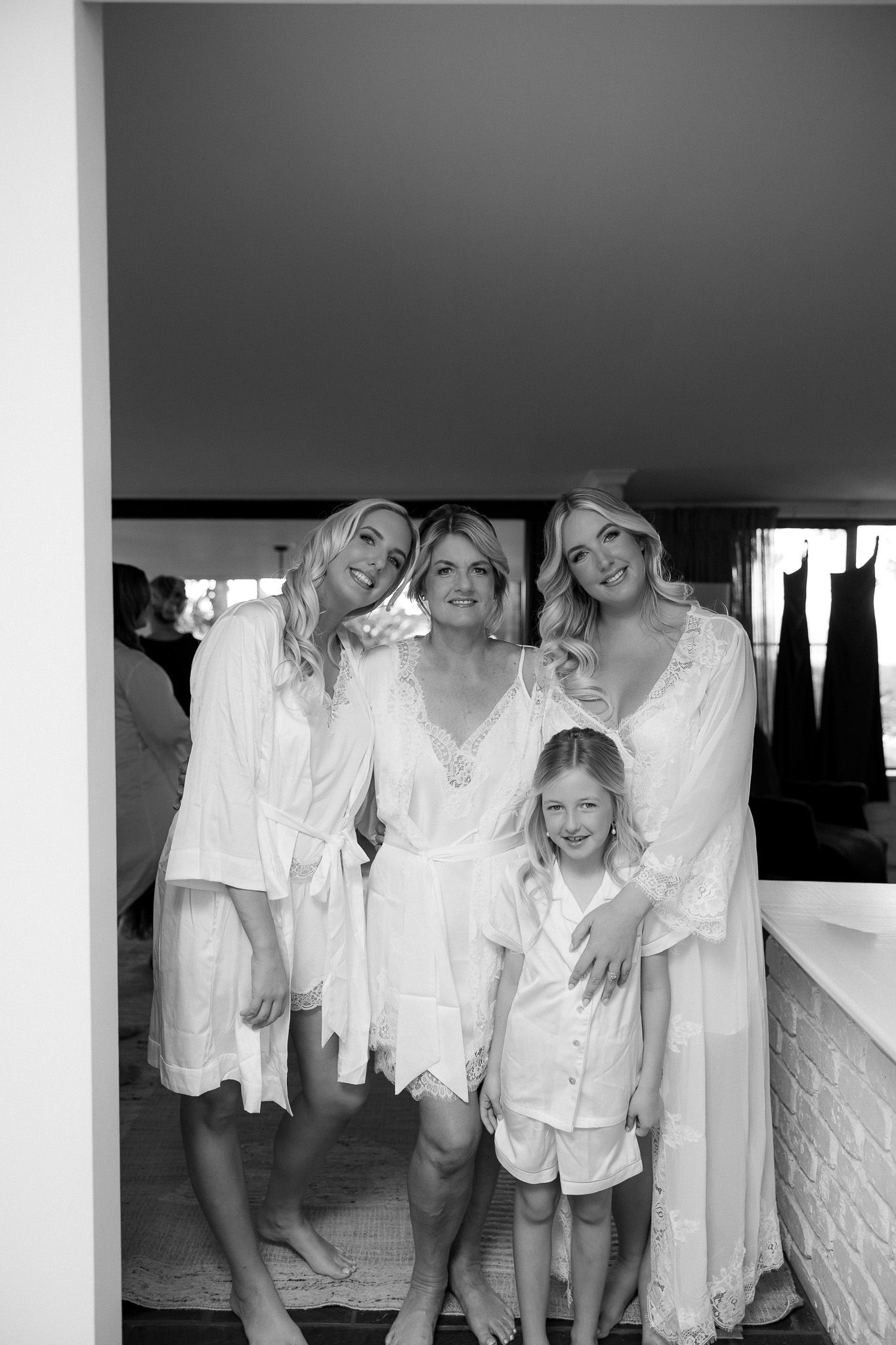 Bride and bridesmaids in white robes smile with young flower girl during wedding morning preparations.