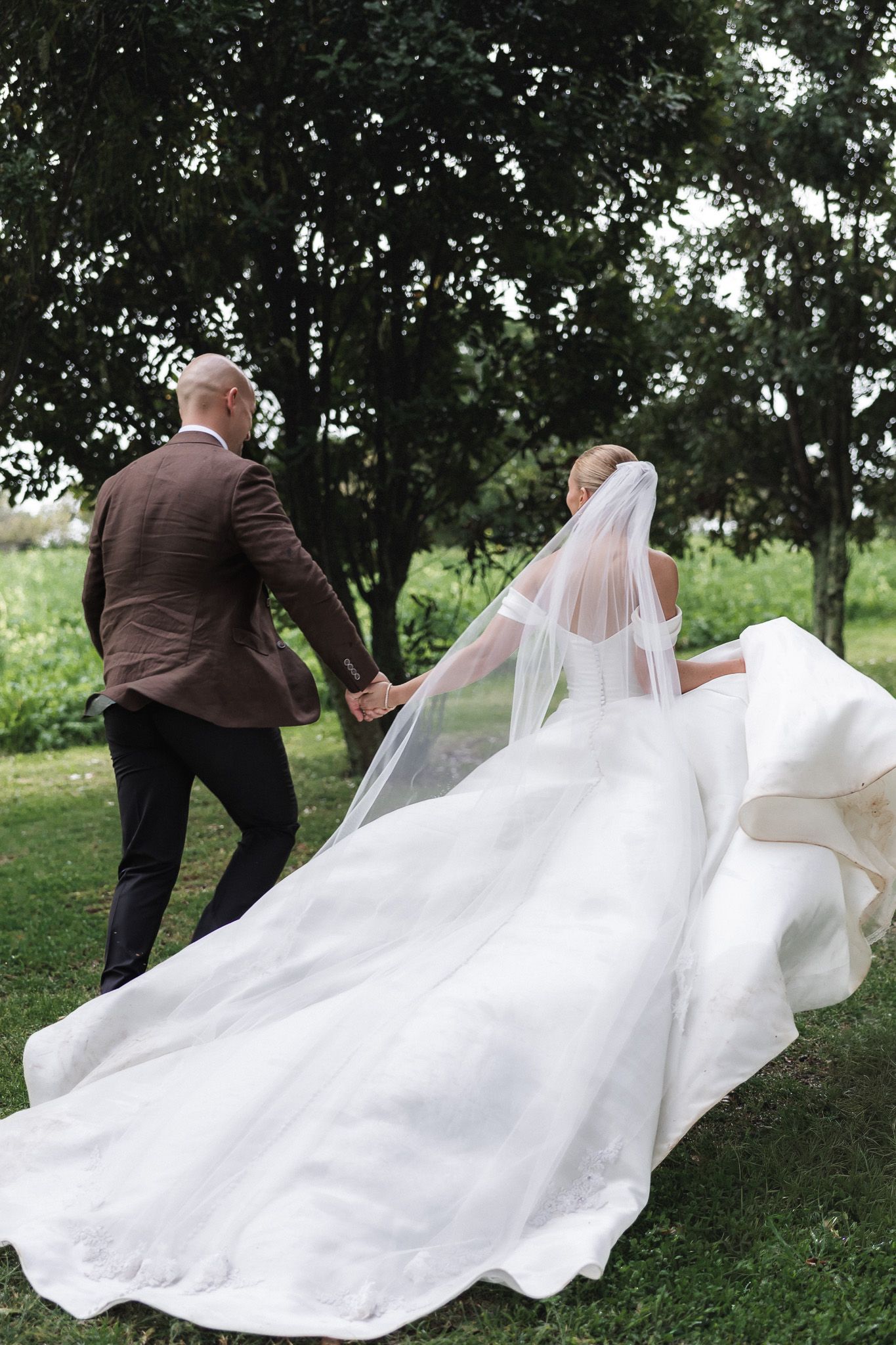 Bride and groom walking hand in hand through lush green garden, veil flowing.