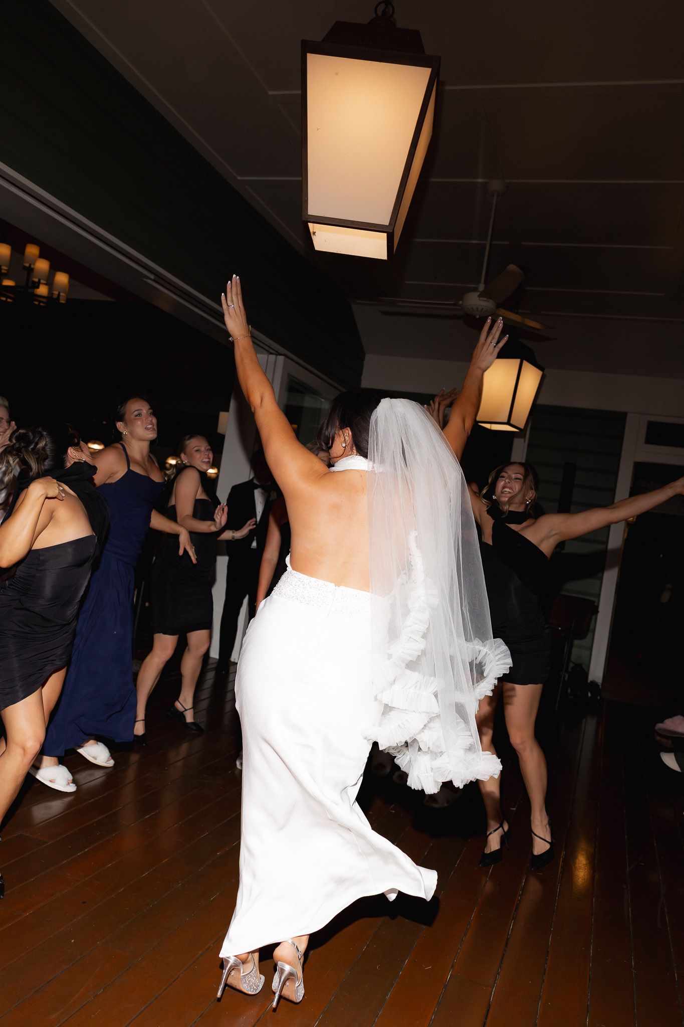 Bride in white gown and veil dancing joyfully with guests at indoor wedding reception under lantern lighting.