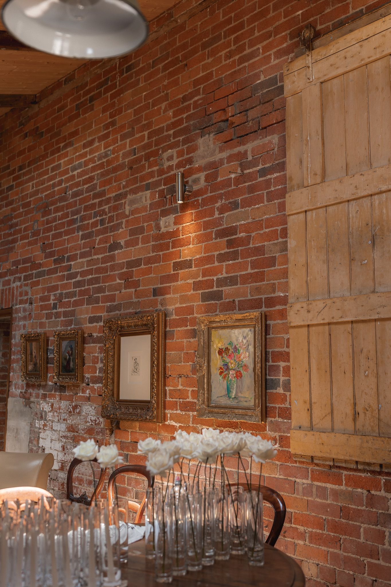 Rustic brick wall wedding venue with white tulips in glass vases, gold-framed artwork, and wooden barn door decor.