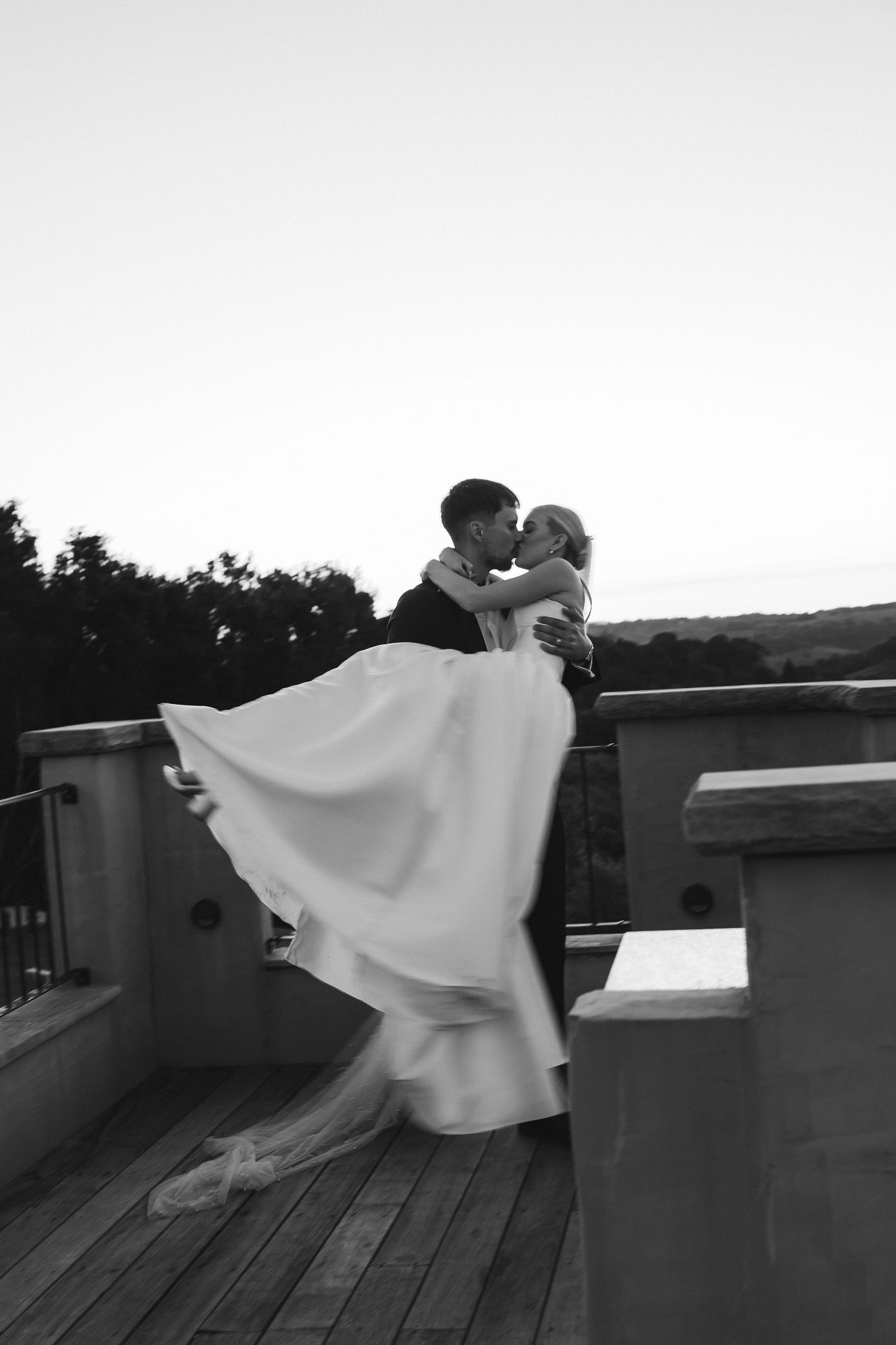 Black and white photo of groom lifting bride in flowing white dress on outdoor terrace at dusk.