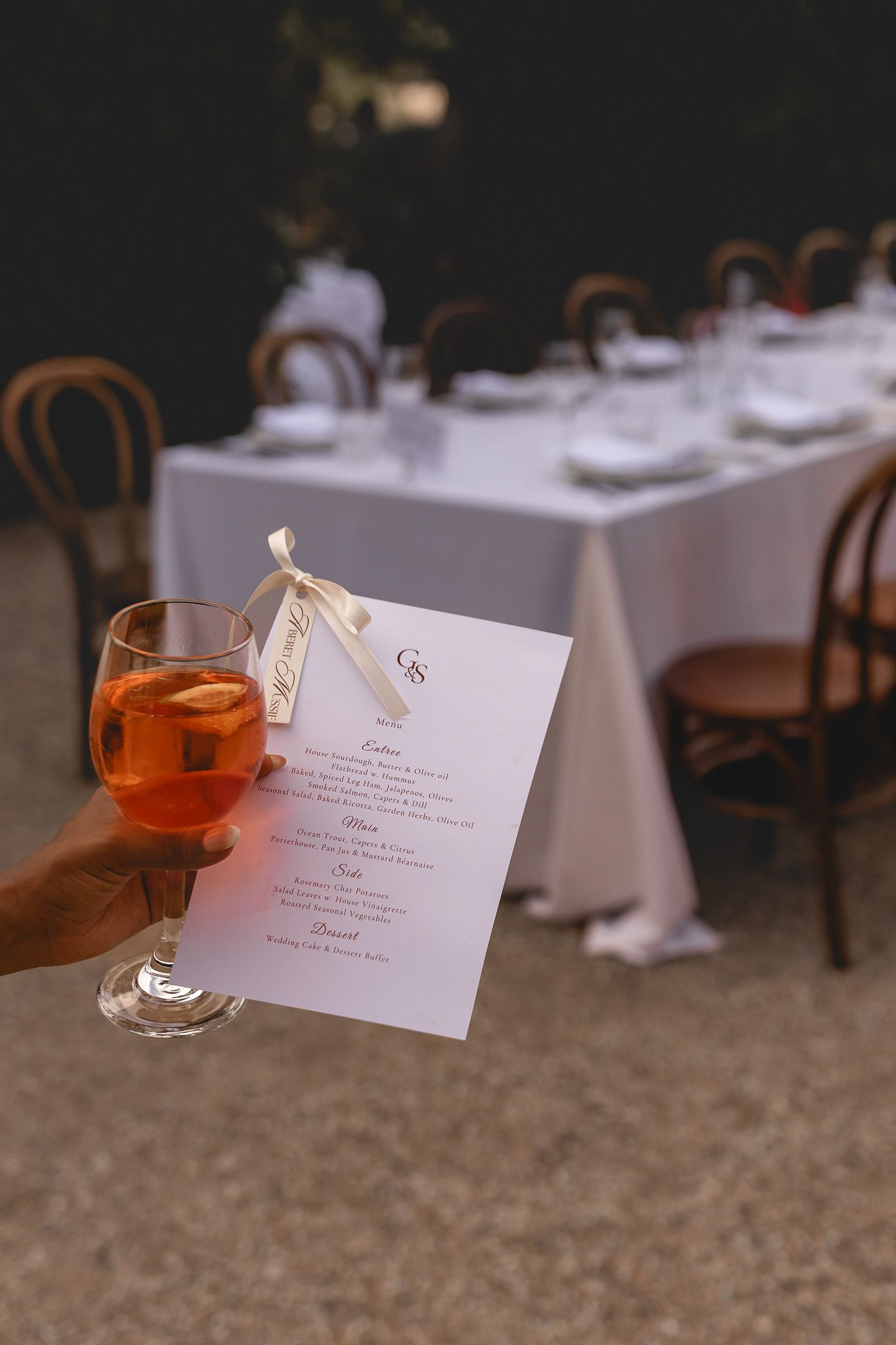 Hand holding an Aperol Spritz cocktail and wedding menu card at an elegant outdoor reception dinner.