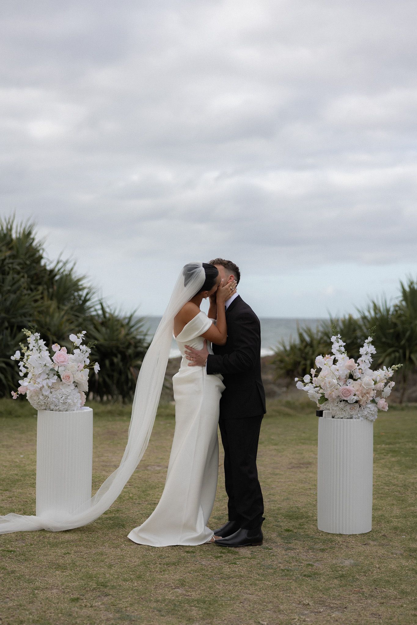 Bride and groom share first kiss at outdoor beachside wedding ceremony with floral arrangements.