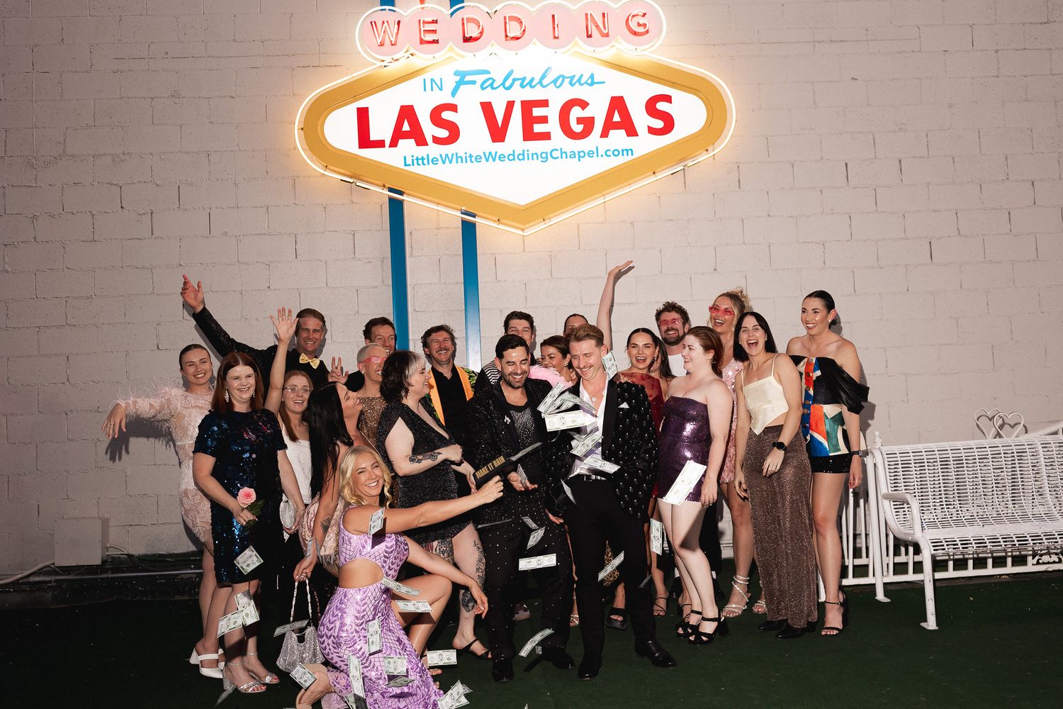 Large wedding party celebrating in front of a glowing Las Vegas wedding sign, tossing confetti at night.