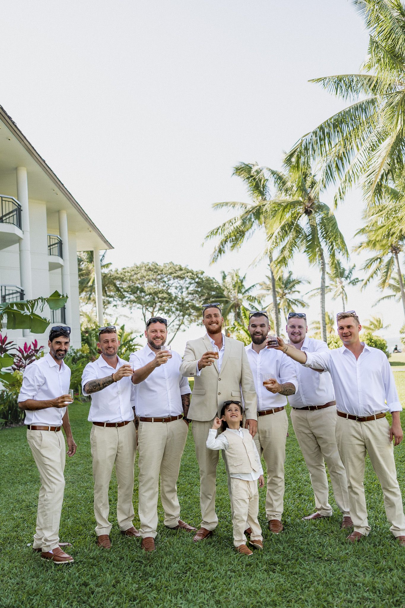 Groom and groomsmen in white shirts and khaki pants pose together on tropical resort lawn.