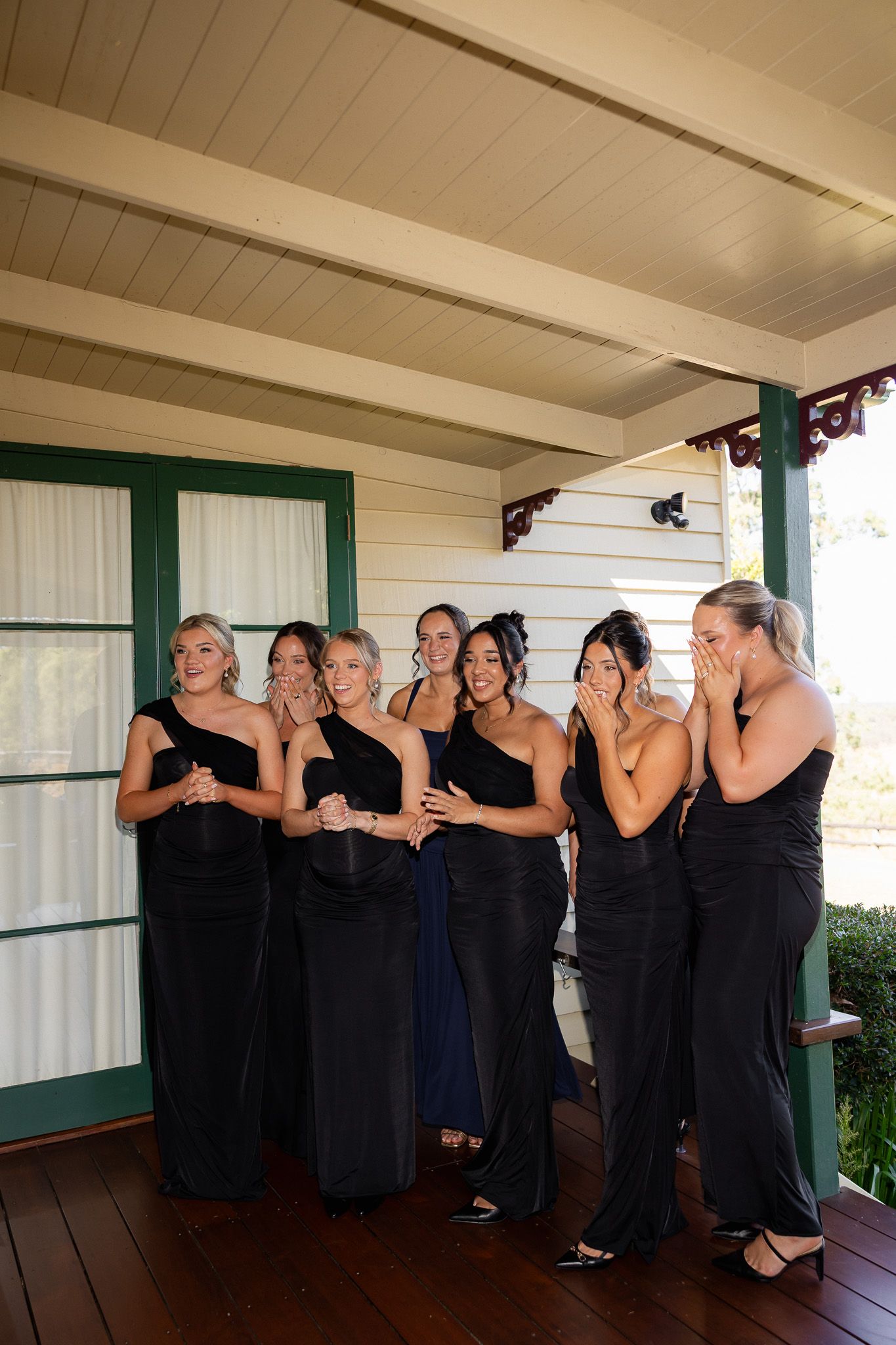 Six bridesmaids in black one-shoulder gowns reacting emotionally on wooden porch before wedding.