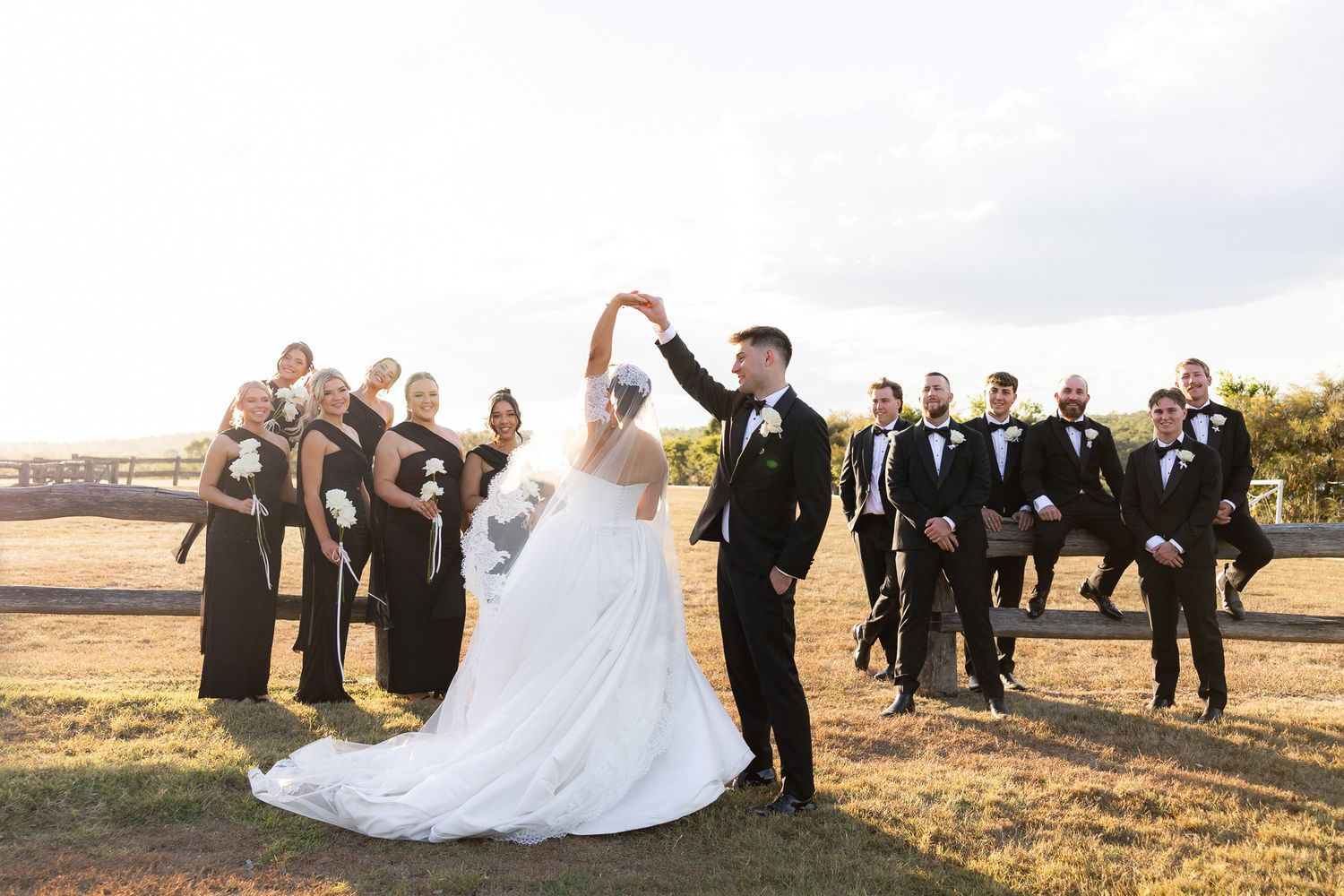 Wedding party walks across field as groom twirls bride in golden hour sunlight.