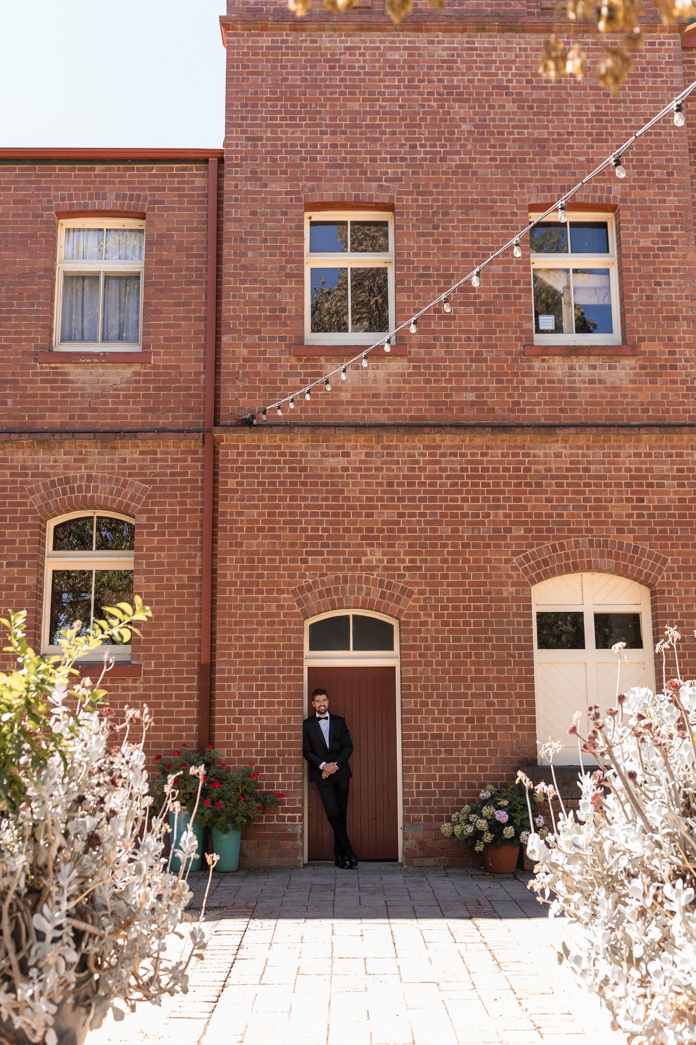 Groom in black tuxedo standing in doorway of rustic red brick building with string lights and garden courtyard.