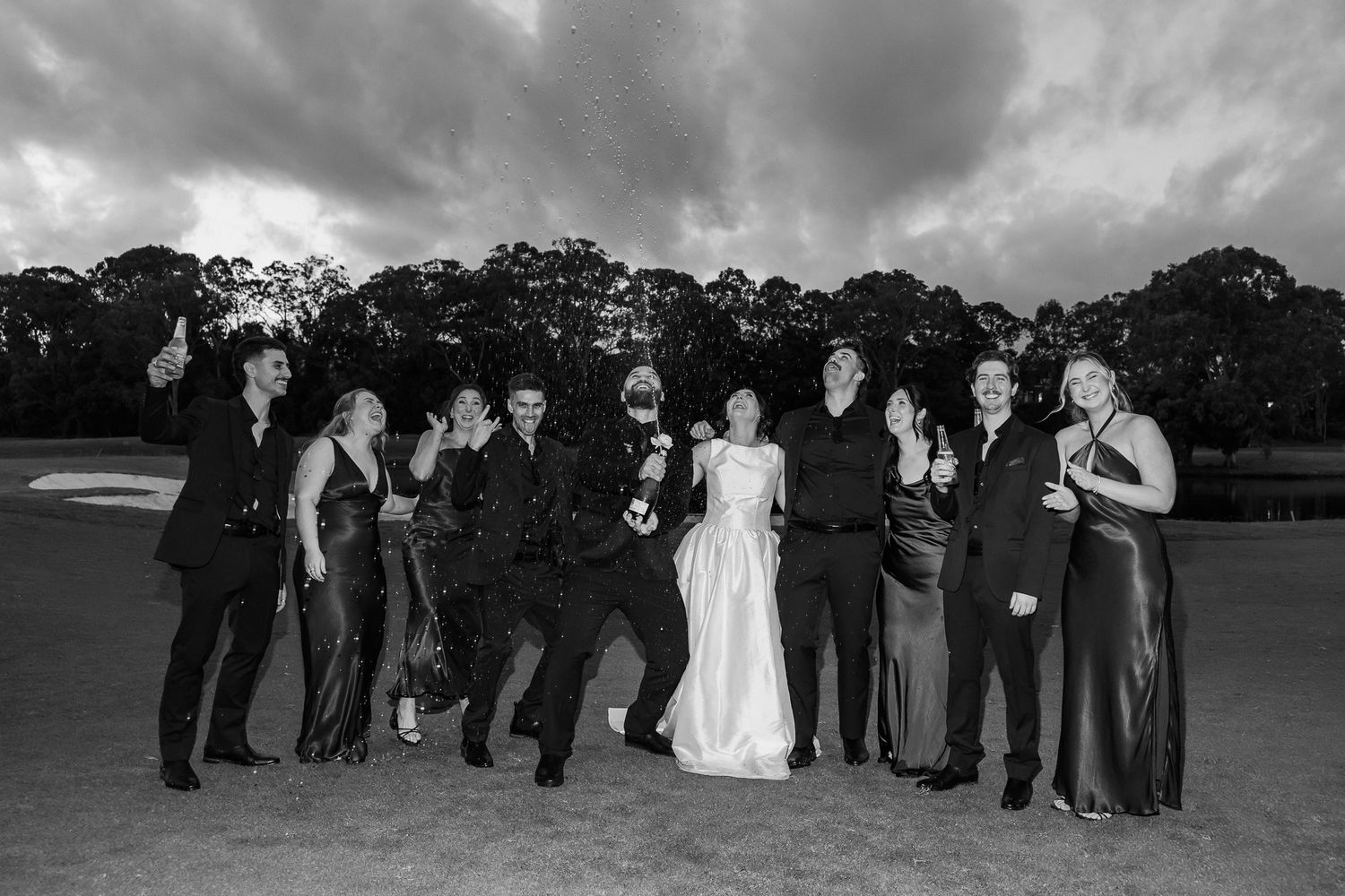 Black and white wedding party photo with bride, groom, and guests celebrating outdoors under dramatic cloudy sky.