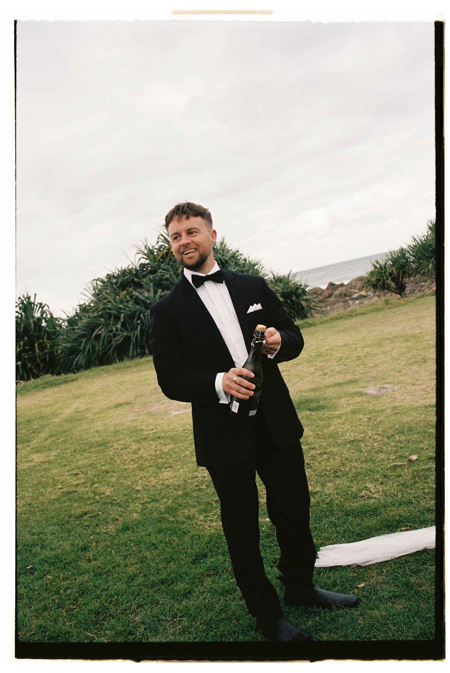 Groom in black tuxedo and bow tie holding champagne bottle on green lawn at outdoor wedding.