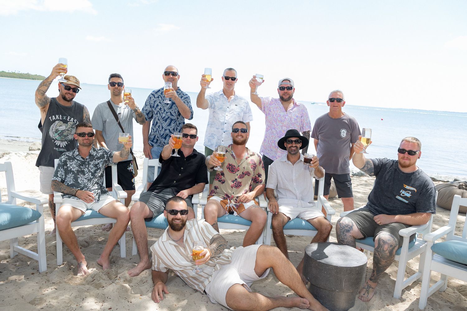 Group of men celebrating on a tropical beach, raising drinks and smiling, with white chairs and ocean in background.