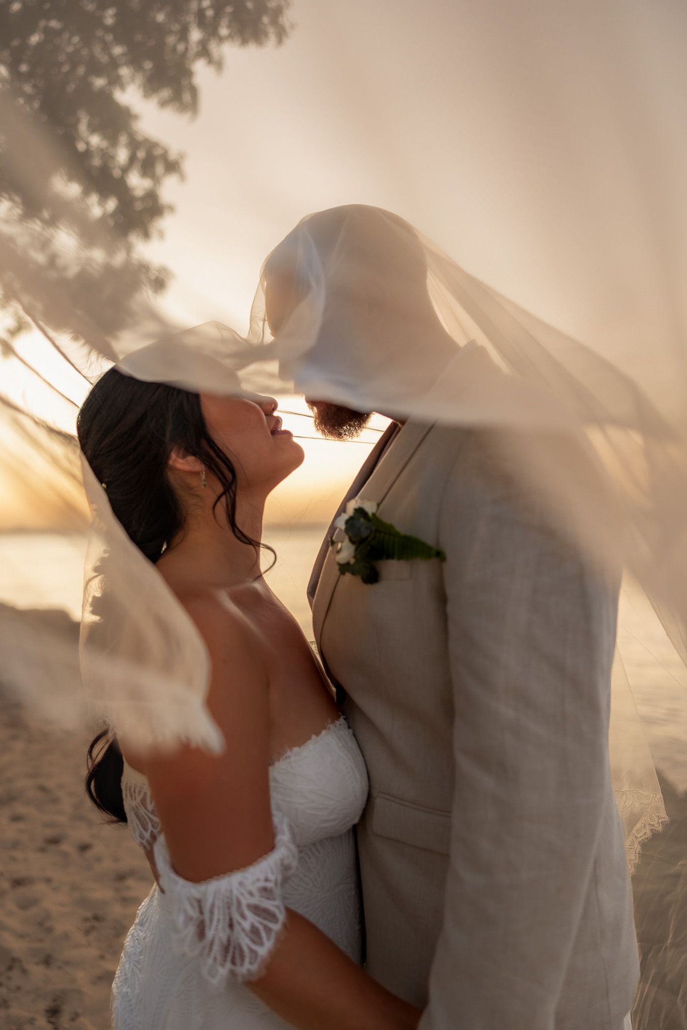 Bride and groom sharing intimate moment under flowing veil on beach at golden sunset.
