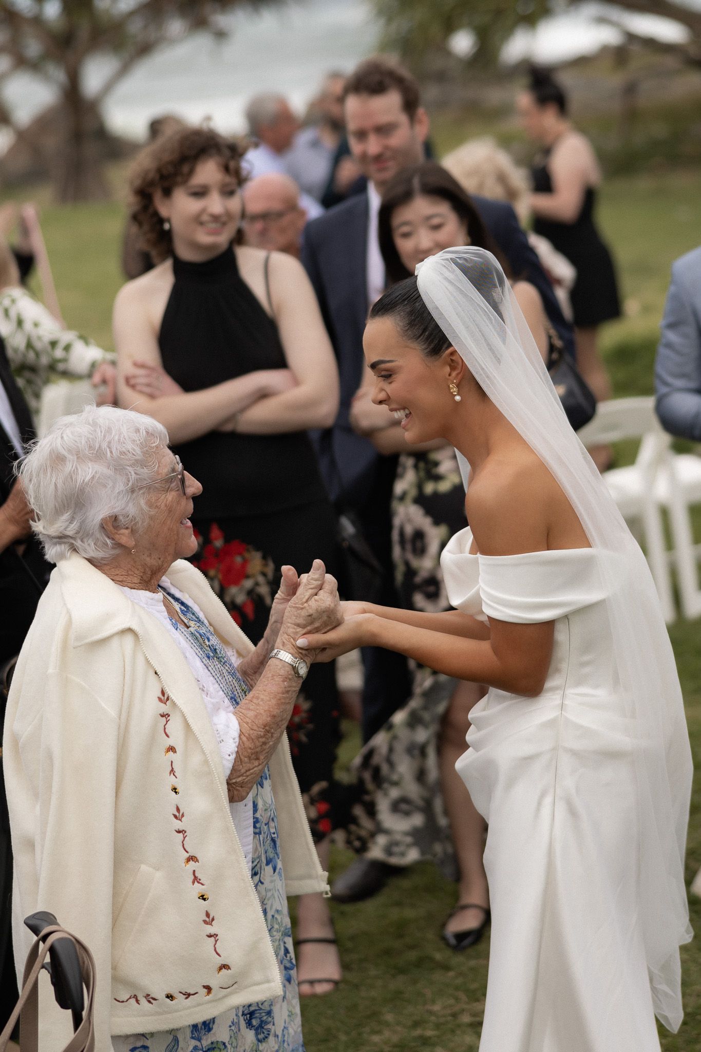 Bride in white gown and veil holds hands with elderly woman at outdoor wedding ceremony.