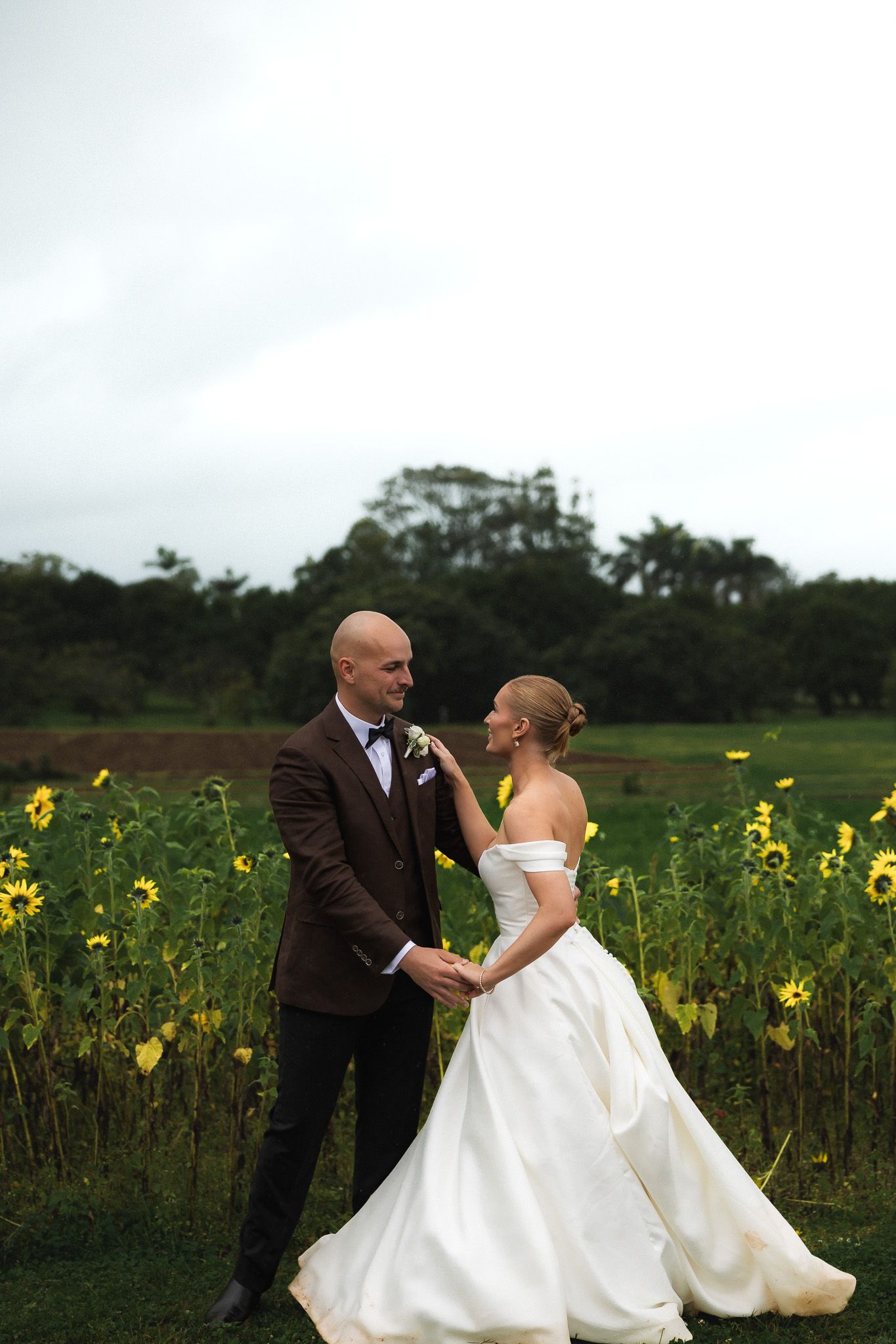 Wedding couple holding hands in sunflower field, bride in off-shoulder gown, groom in brown suit.