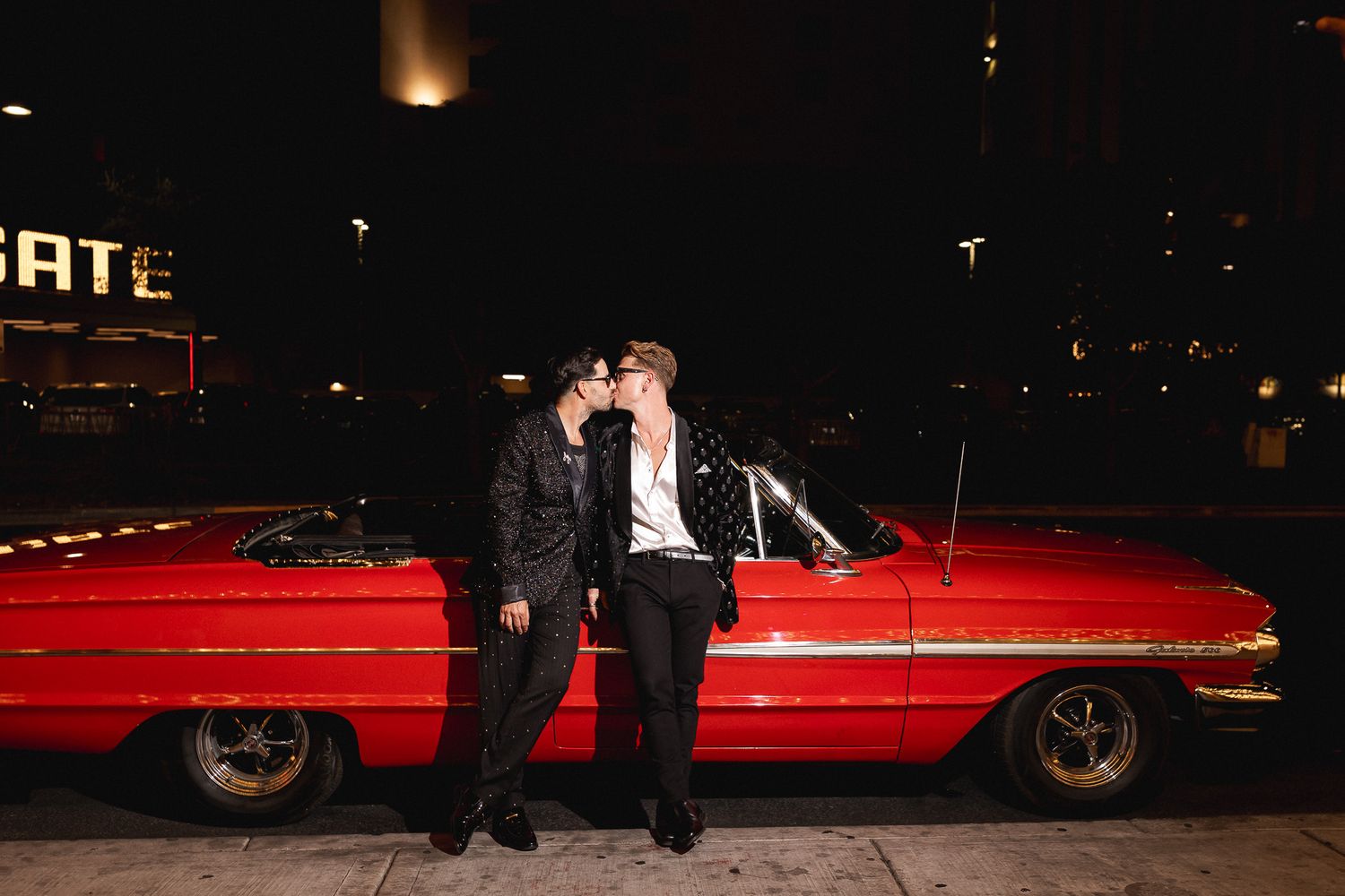 Two grooms sharing a kiss beside a vintage red convertible car on a Las Vegas street at night.