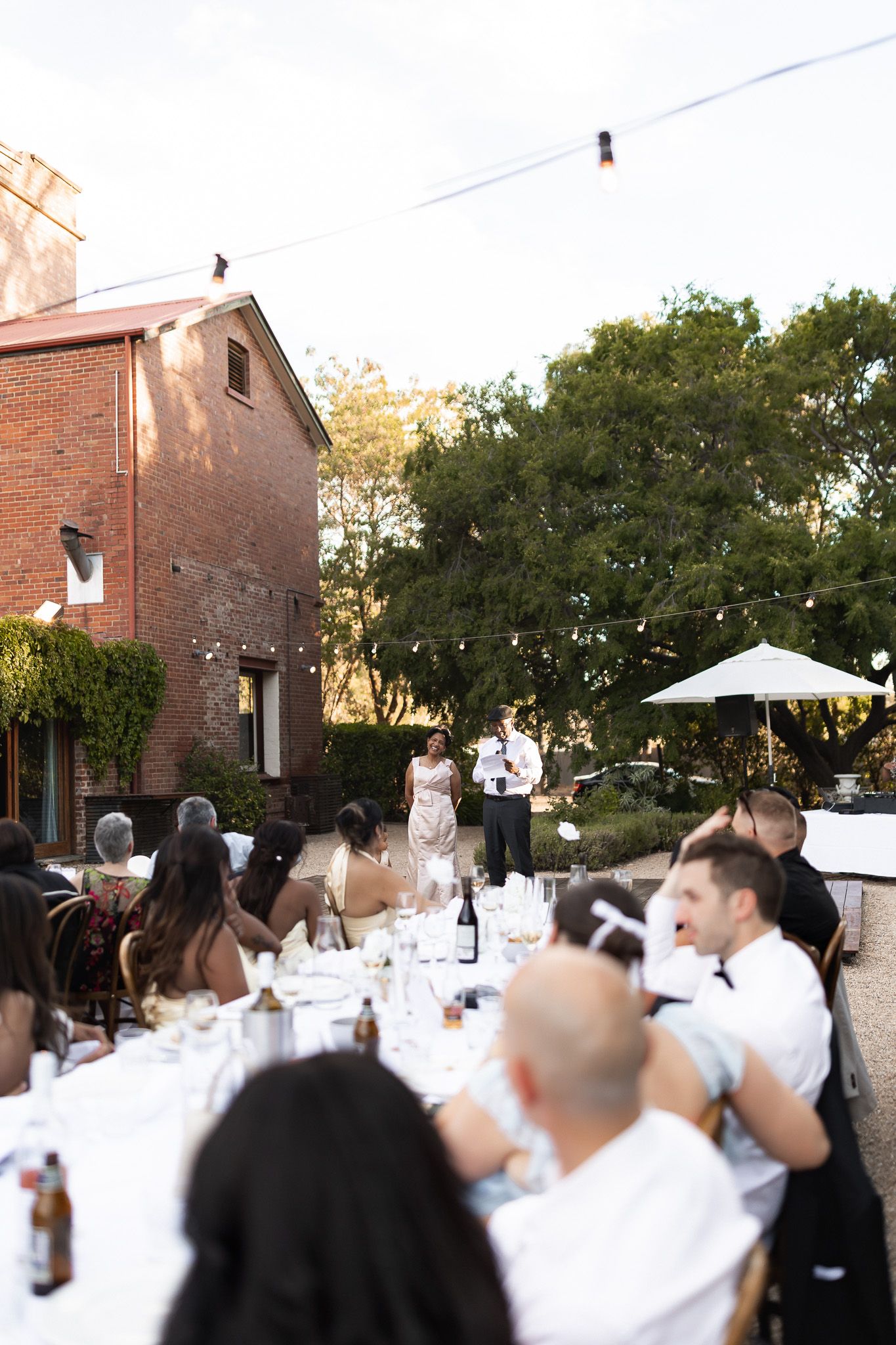 Bride and groom giving speeches at an outdoor wedding reception with string lights and brick building backdrop.