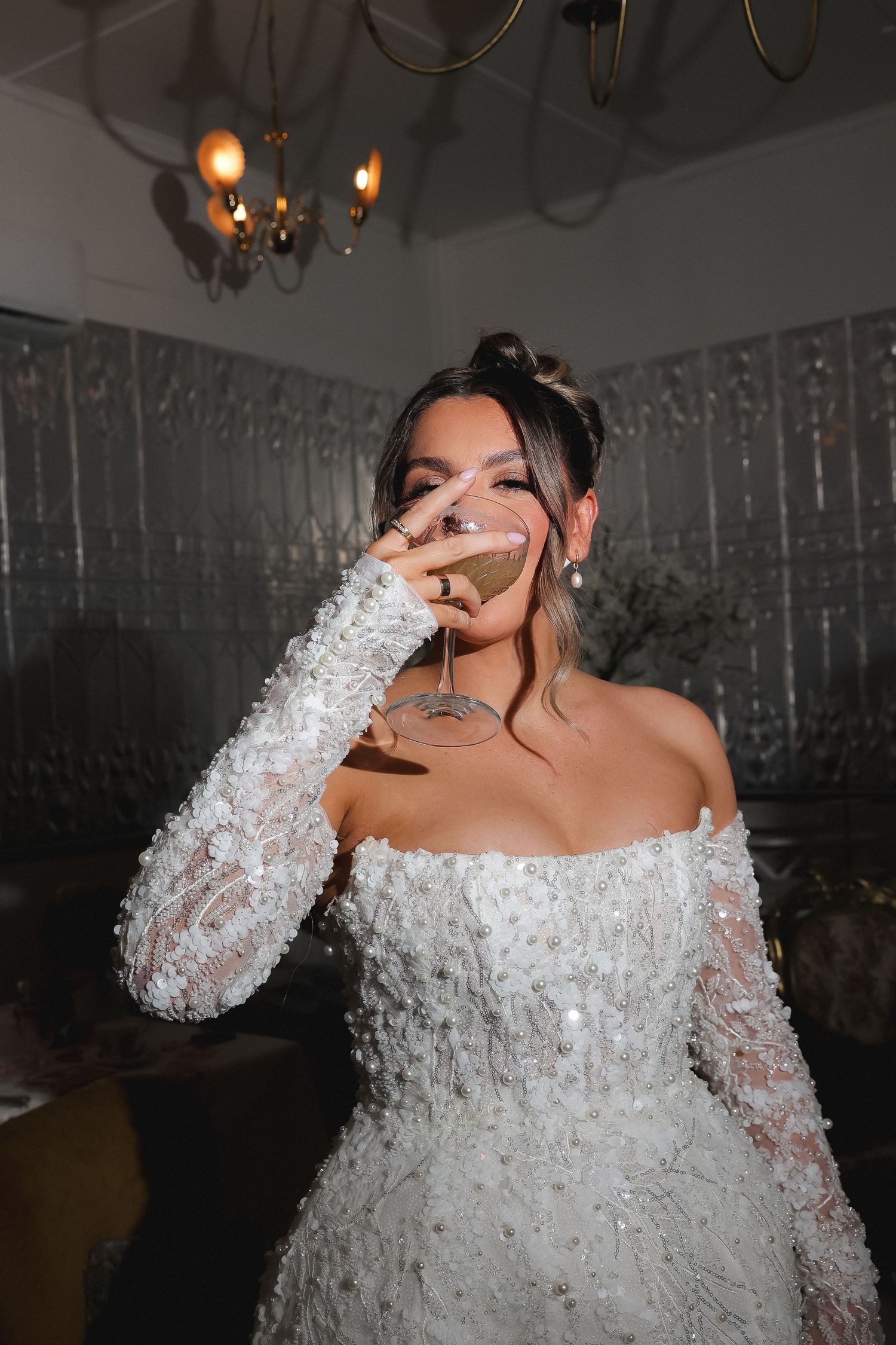Bride in beaded off-shoulder gown drinking champagne at wedding reception under chandelier.