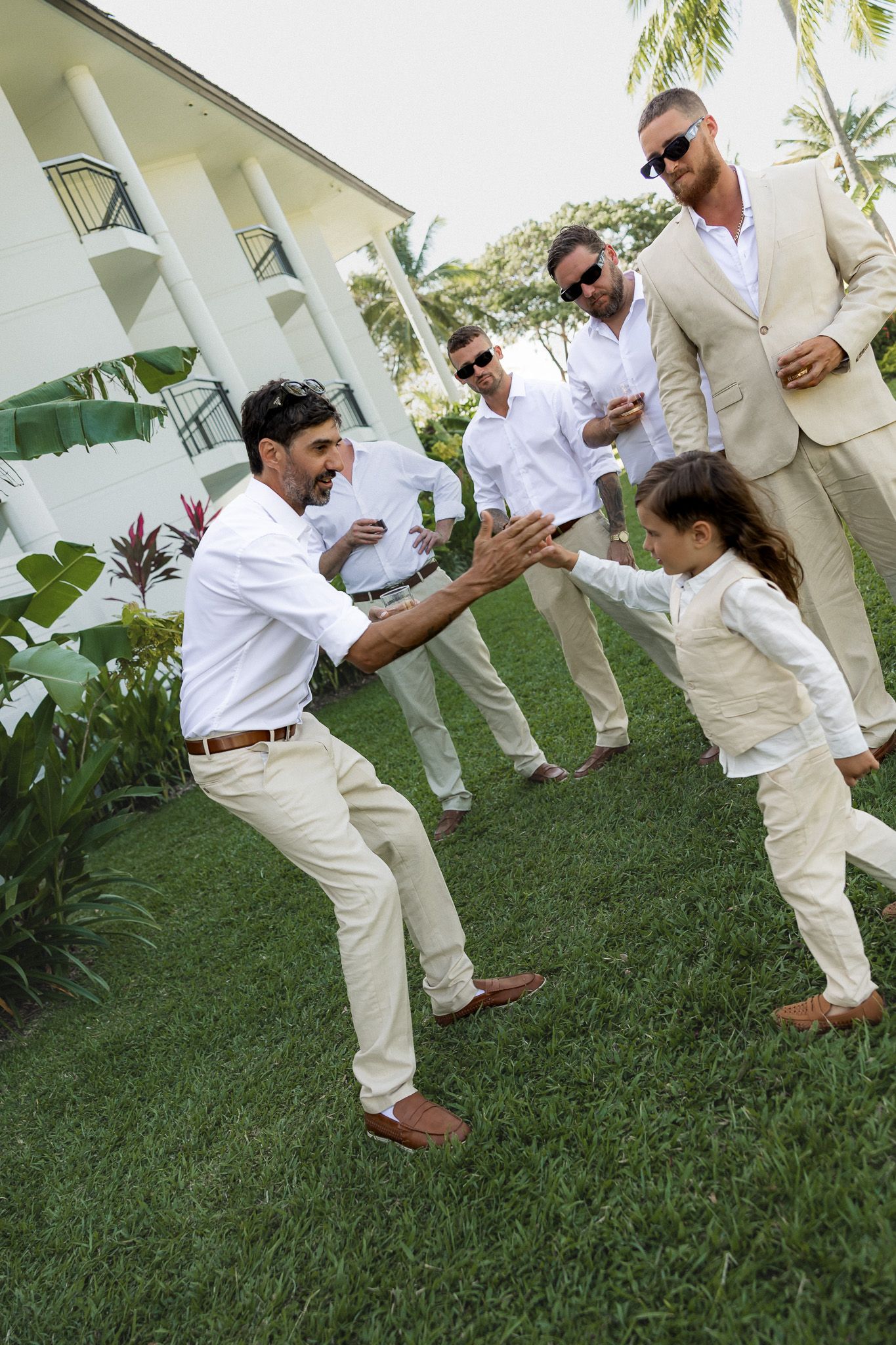 Men in white wedding attire high-five a young child on green lawn at tropical venue.