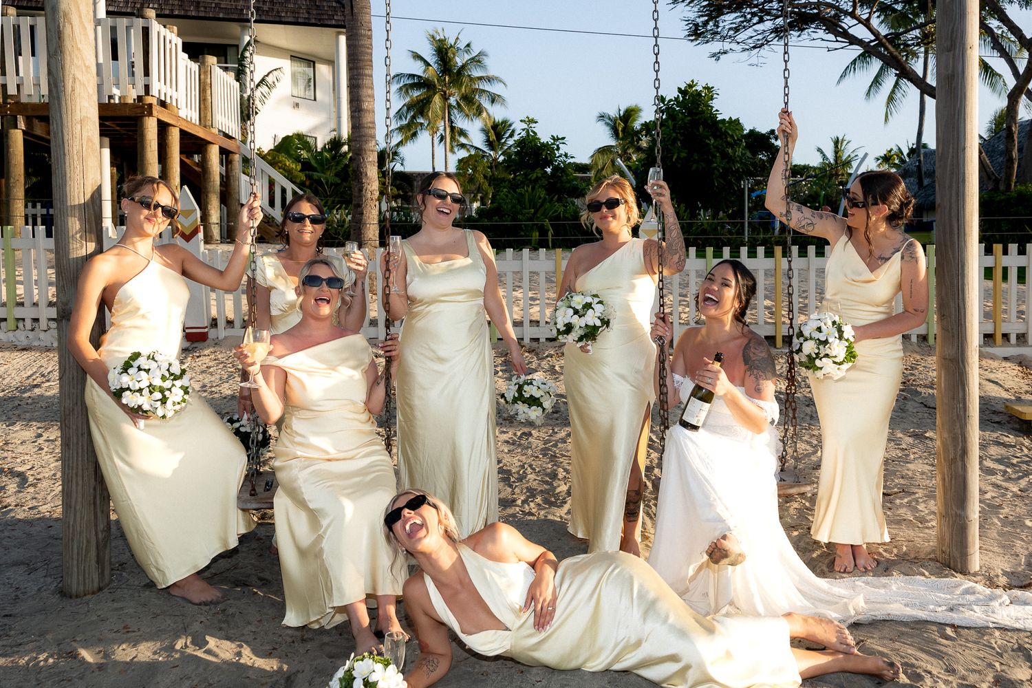 Bridesmaids in cream dresses pose playfully on a sandy beach with bouquets at a destination wedding.
