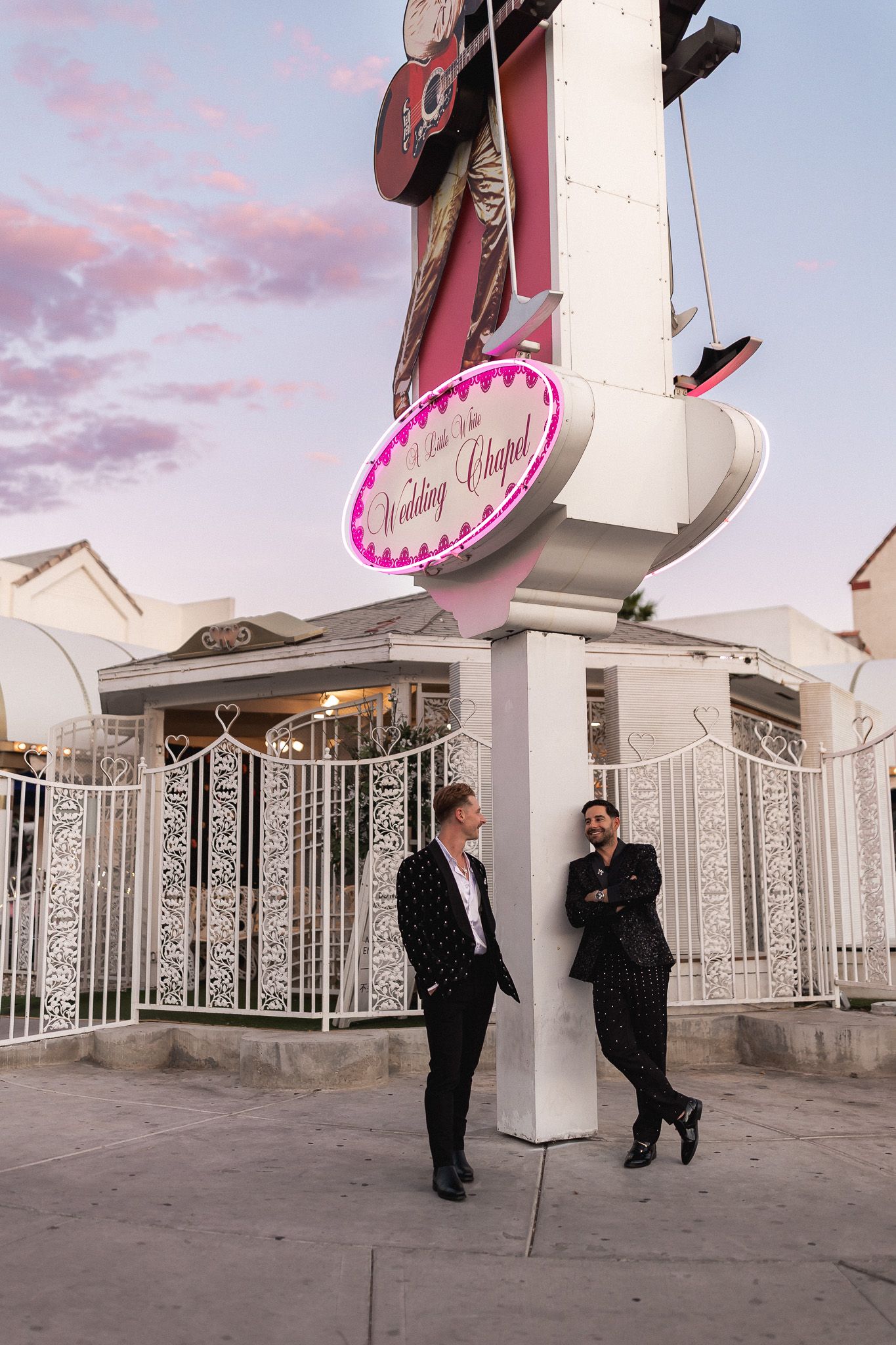 Two grooms in black suits pose outside A Little White Wedding Chapel in Las Vegas at dusk.