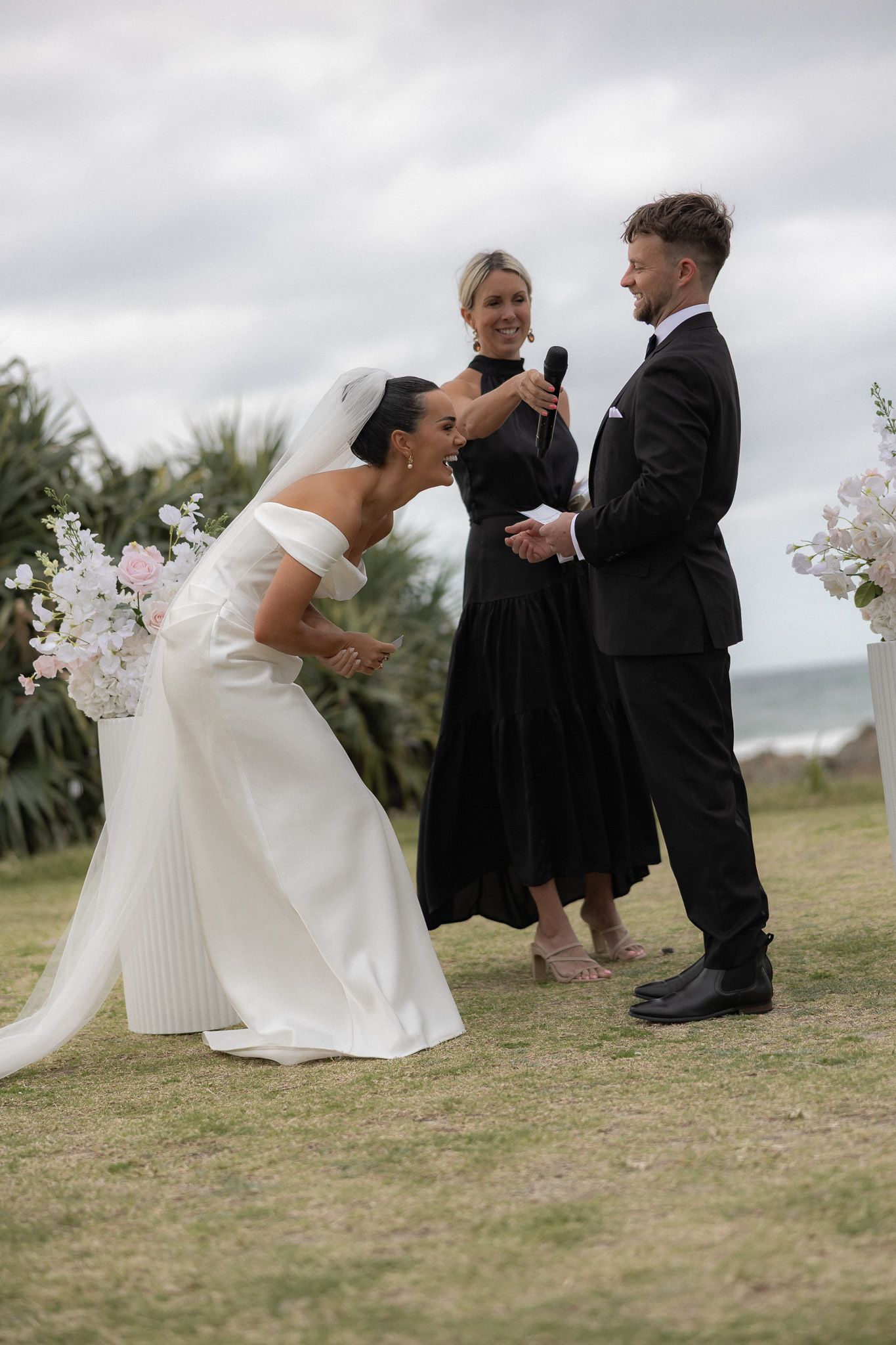 Bride laughing joyfully during outdoor wedding ceremony by the ocean with groom and officiant.