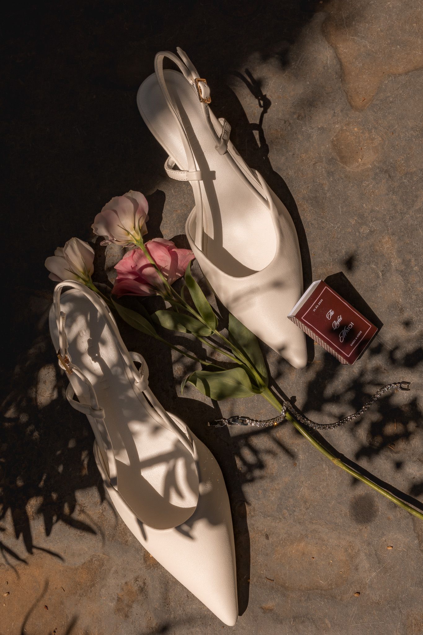 White slingback heels with pink roses and a jewelry box arranged on a stone surface in warm sunlight.