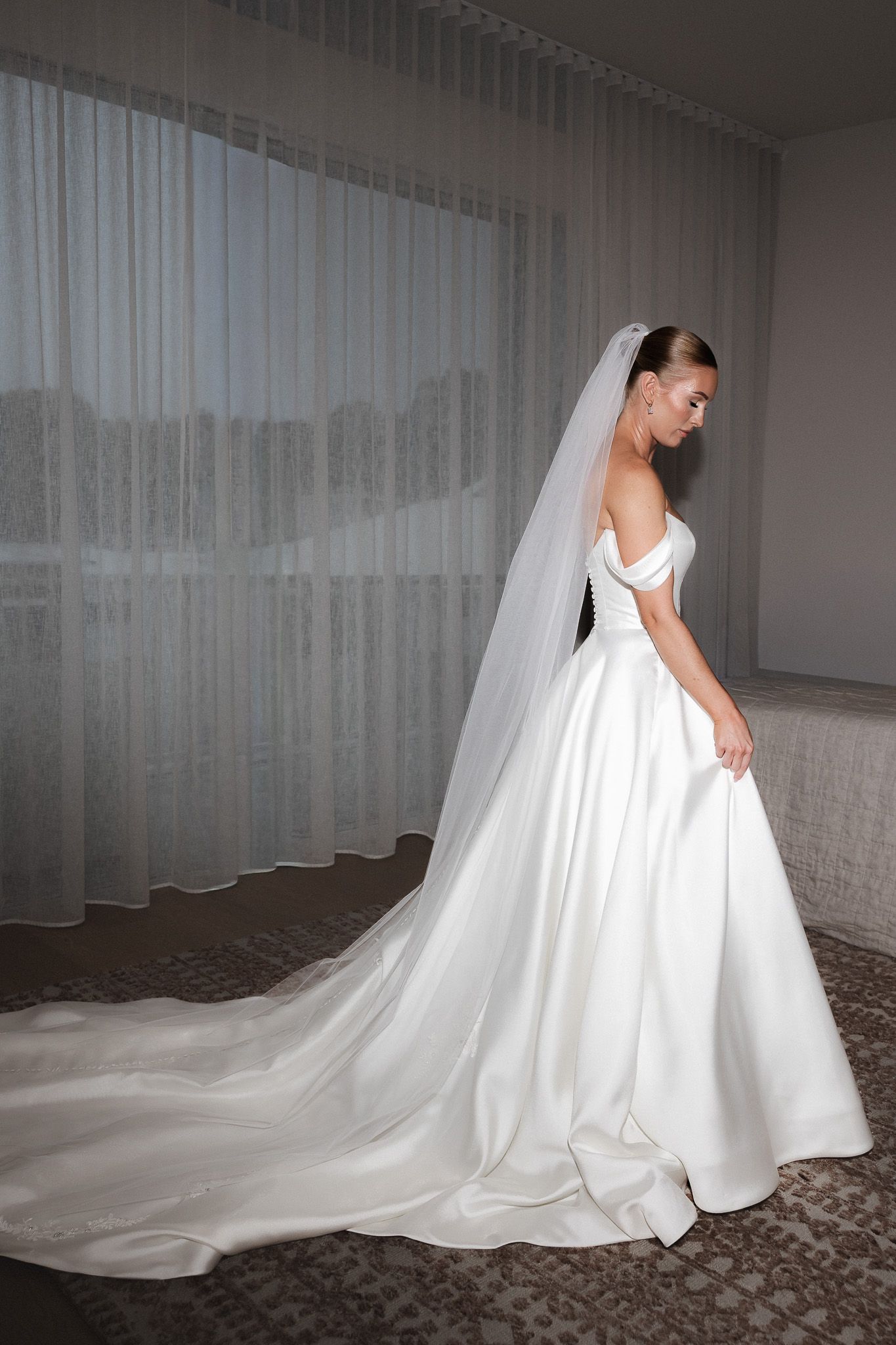 Elegant bride in off-shoulder ballgown with long cathedral veil standing by window.