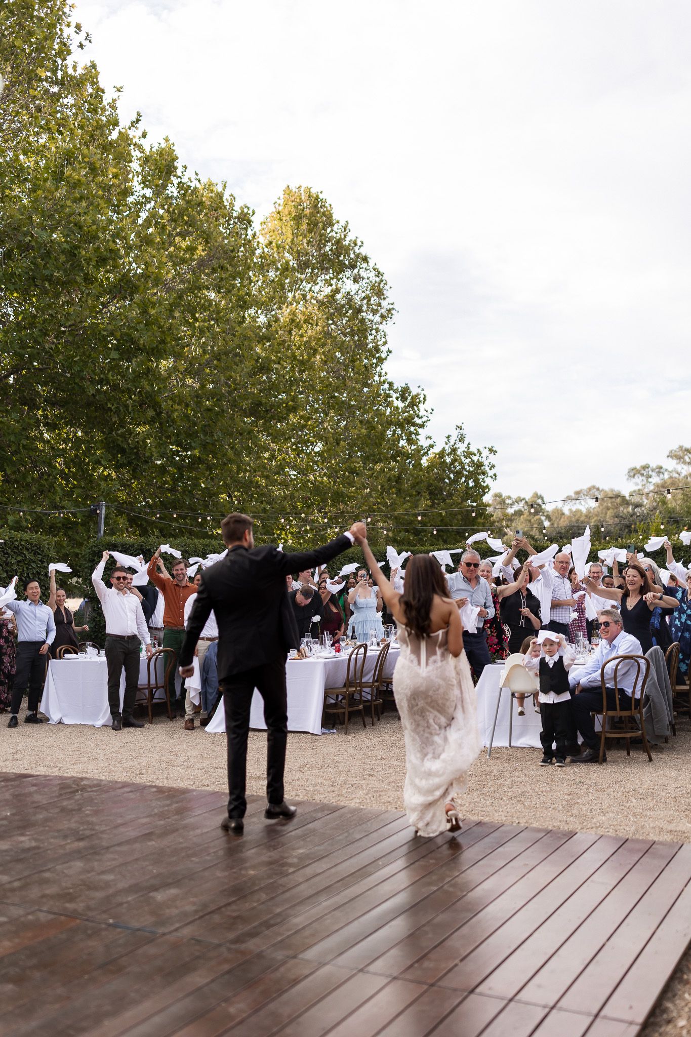 Bride and groom share first dance on outdoor wooden stage as wedding guests wave white napkins in celebration.