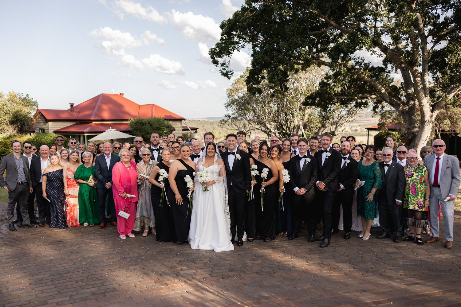 Large wedding party group photo outdoors with bride, groom, and guests at rustic venue.