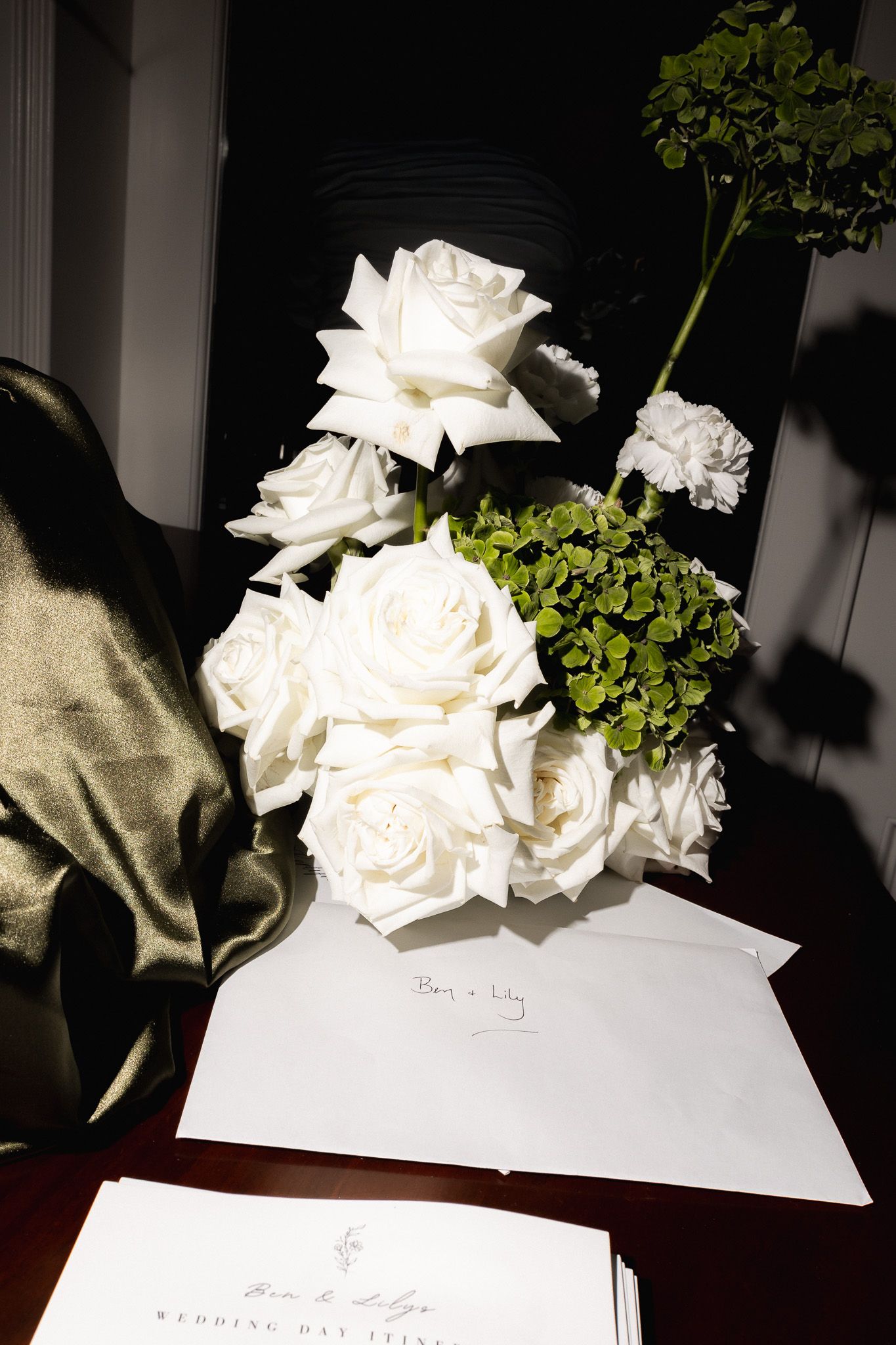 Elegant white roses and green hydrangea wedding bouquet beside handwritten wedding day itinerary cards on dark wood.
