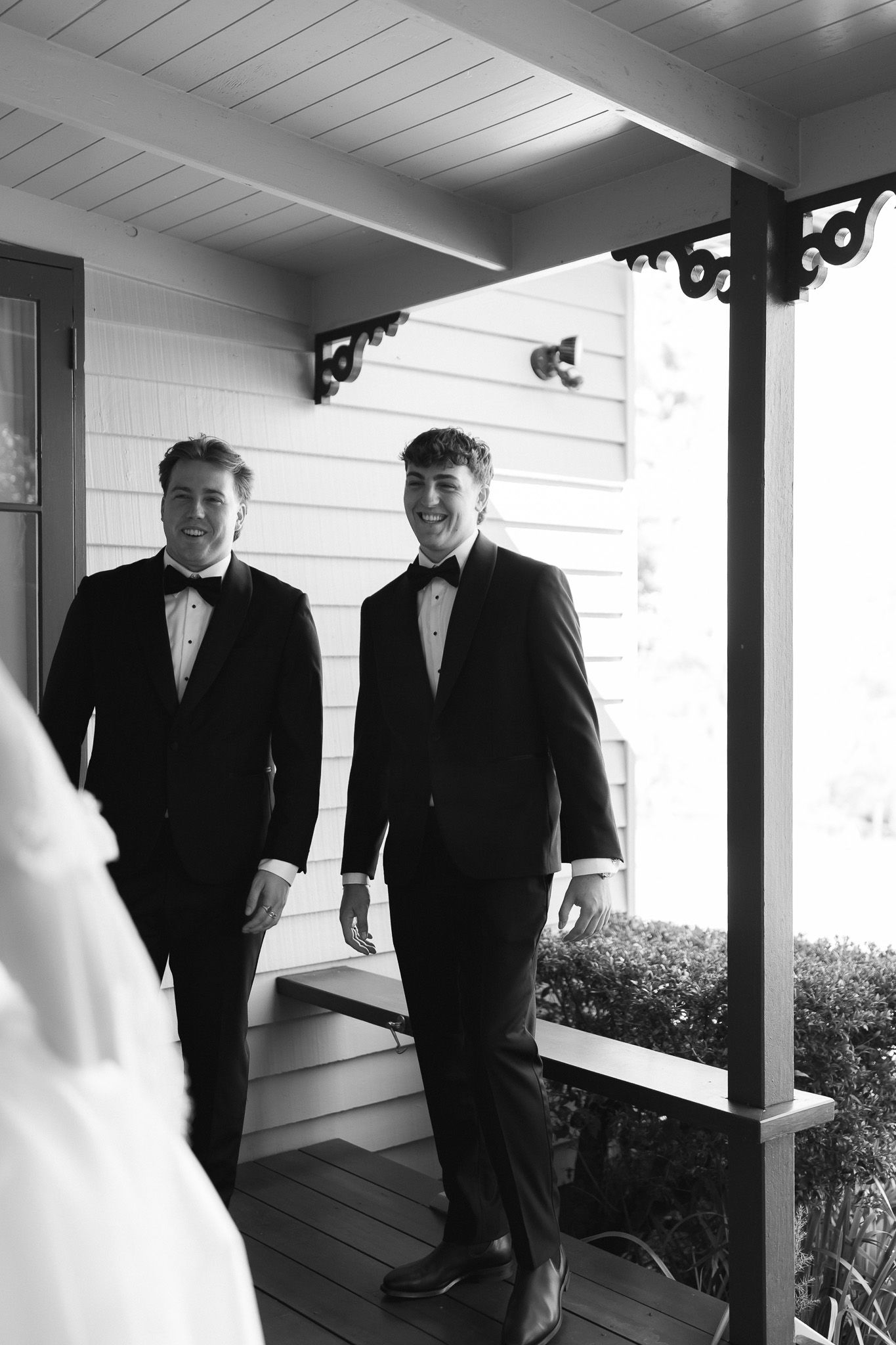 Two groomsmen in black tuxedos smiling on a porch in a black and white wedding photo.