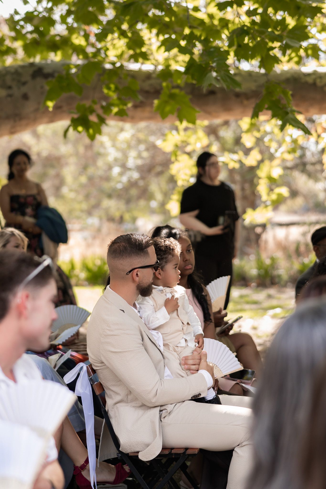 Guest in beige suit holds child during outdoor wedding ceremony seated under green leafy tree canopy.