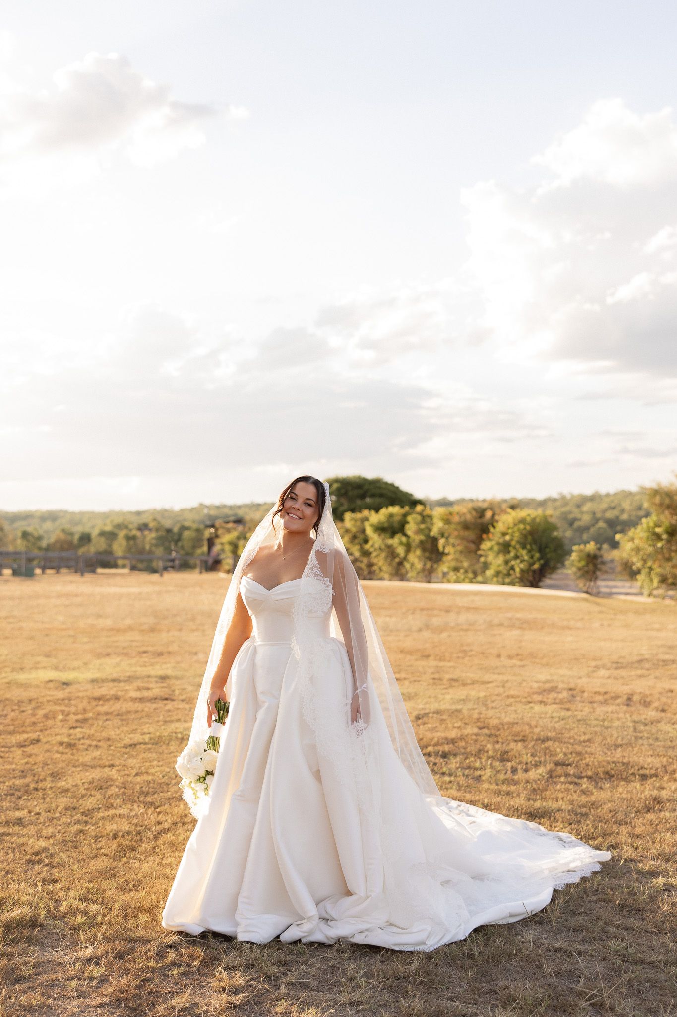 Bride in white ball gown with veil standing in golden field at sunset.
