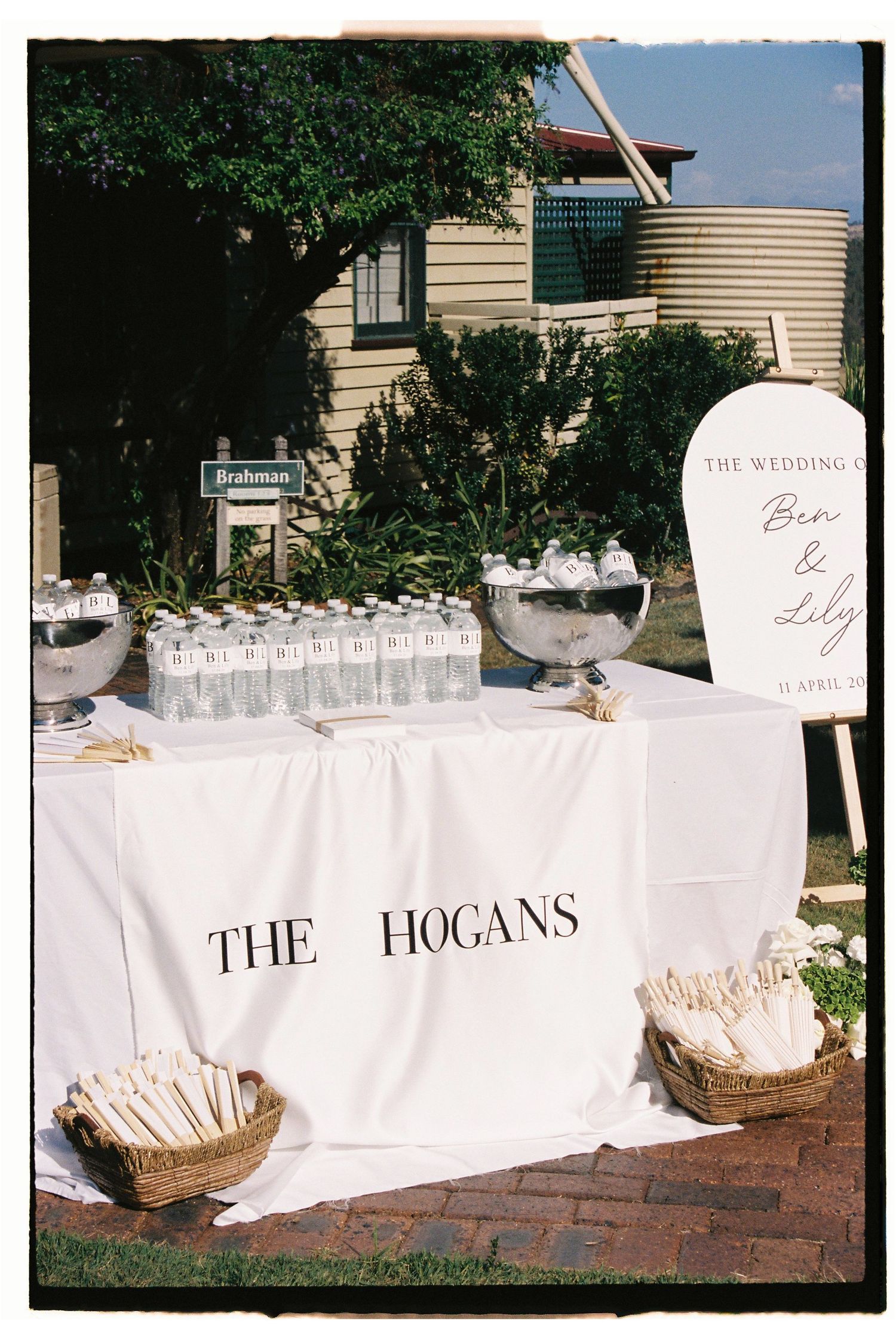 Elegant wedding welcome table with white tablecloth reading The Hogans, featuring water bottles, floral arrangements, and a bridal sign.