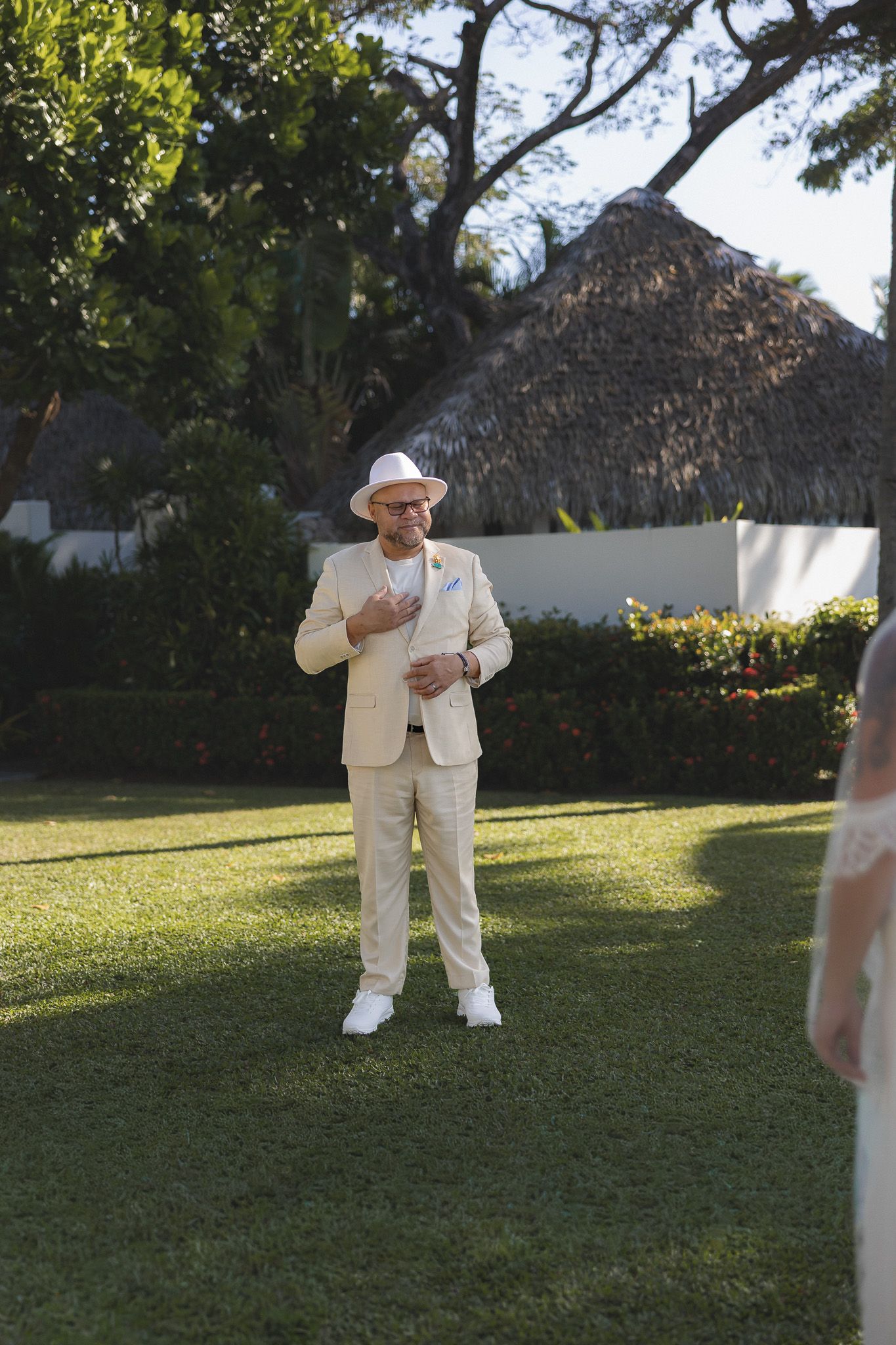 Groom in beige suit and white hat stands on garden lawn during outdoor wedding ceremony at tropical venue.