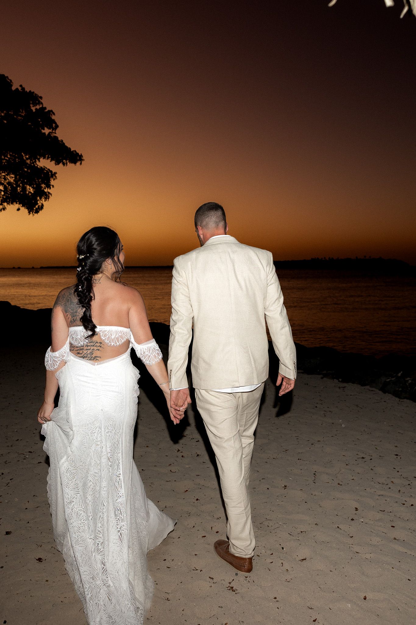 Bride and groom walk hand in hand along beach at dramatic orange sunset after wedding.