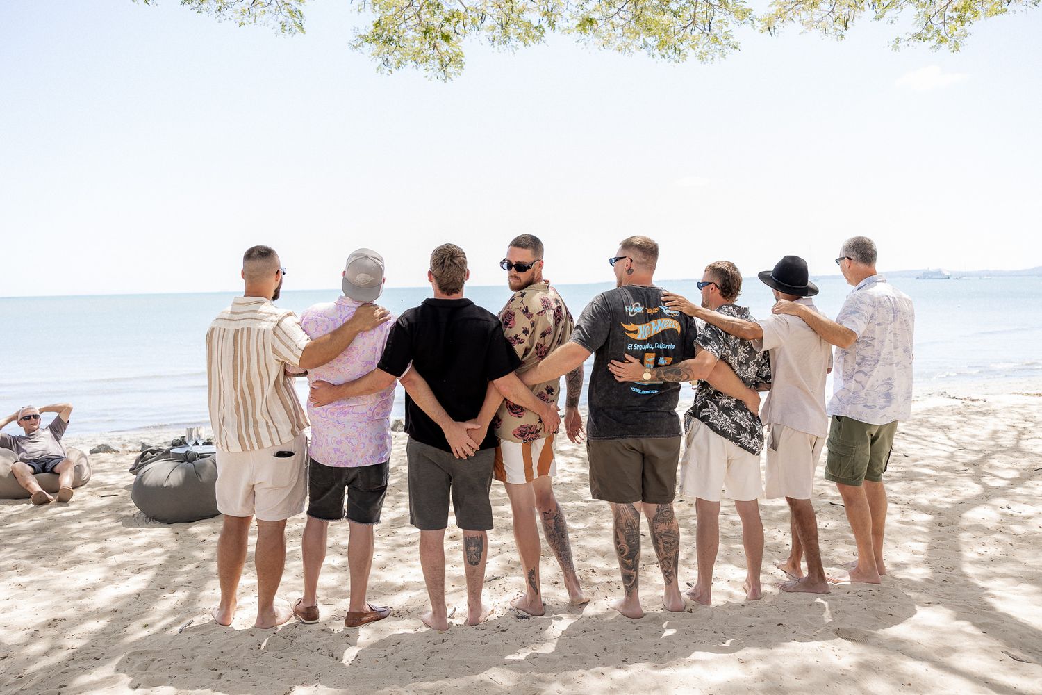 Group of friends standing together on a sandy beach, arms around each other, facing the ocean on a sunny day.