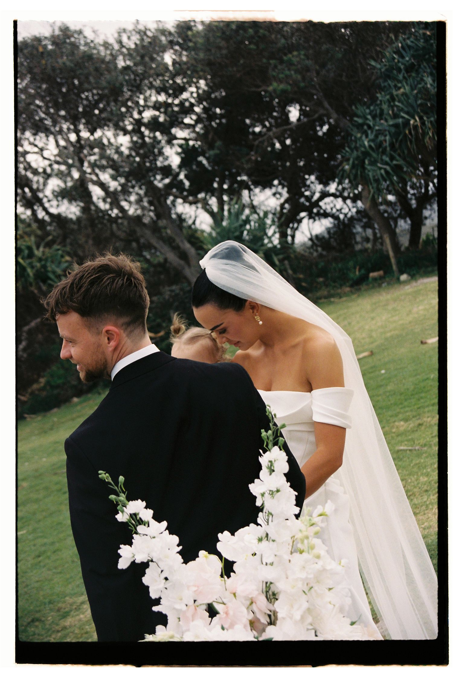 Bride and groom share an intimate moment outdoors, with bride holding white florals wearing off-shoulder gown and veil.