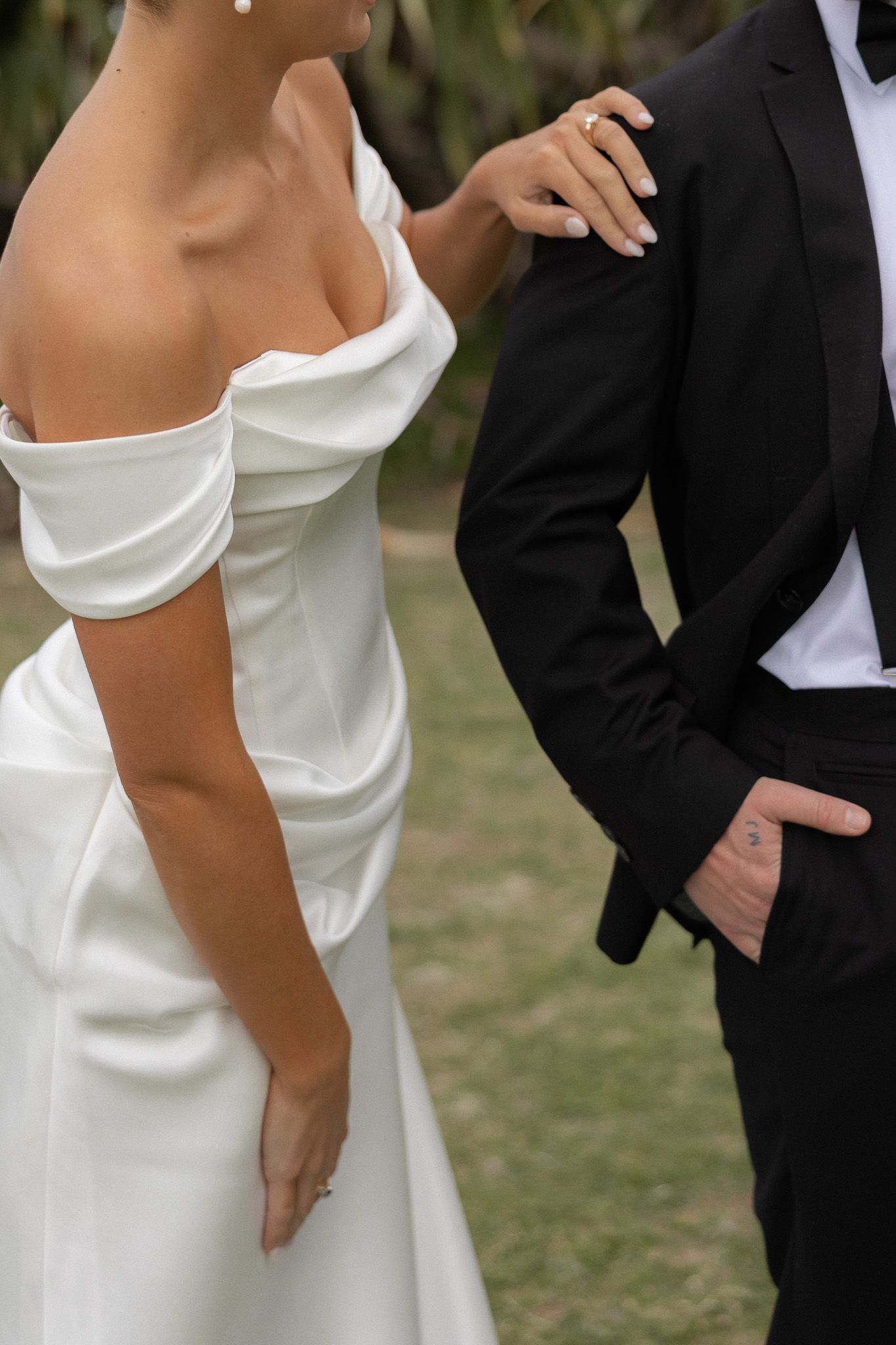Close-up of bride in elegant off-shoulder white satin gown resting hand on grooms black suit jacket.