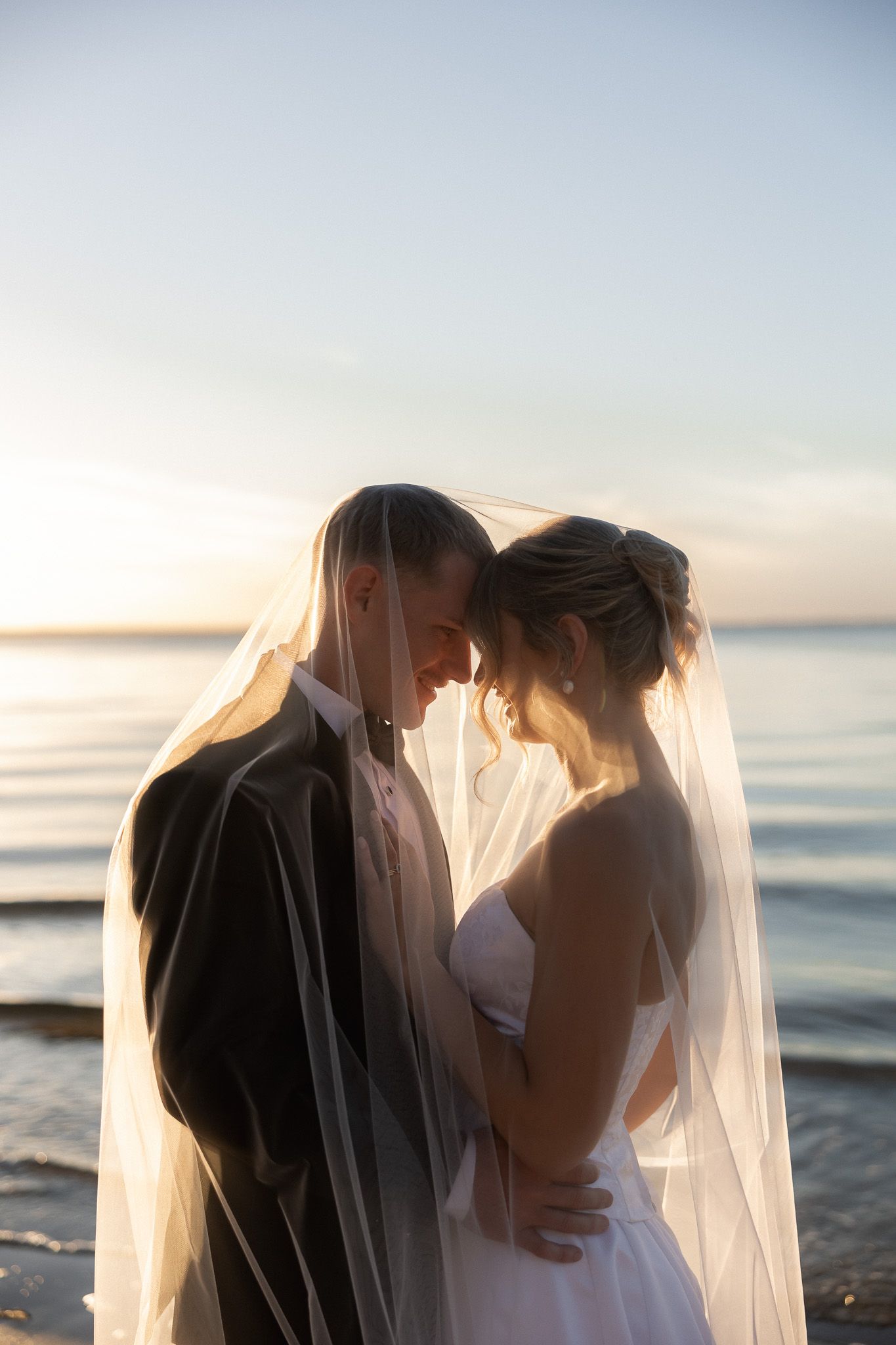 Bride and groom share romantic moment under veil at sunset by the ocean on their wedding day.
