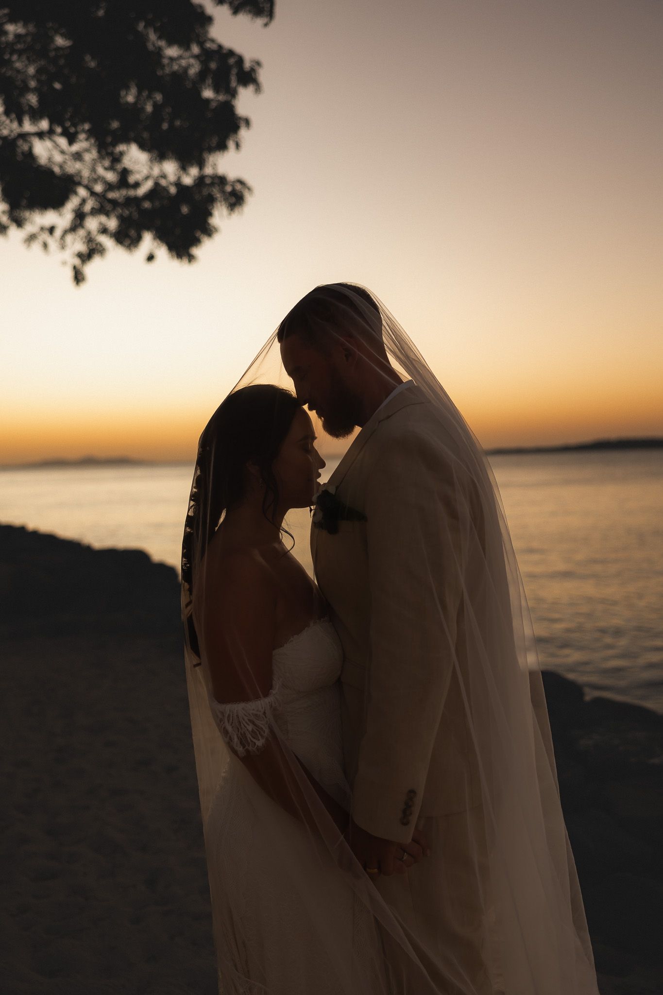Silhouette of bride and groom sharing romantic moment under veil at sunset by the ocean.