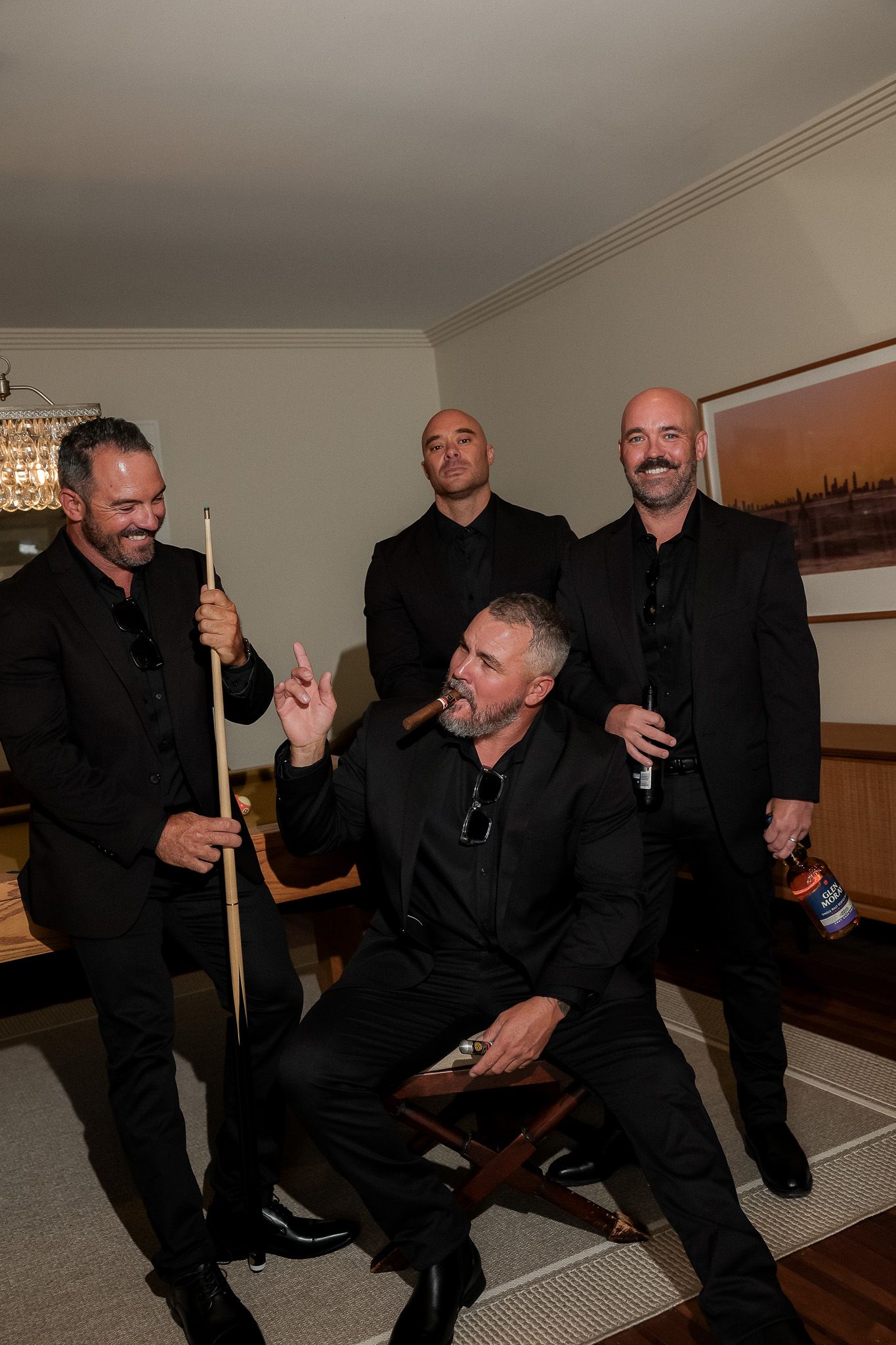 Four men in black suits laughing and posing with pool cue and cigar indoors.