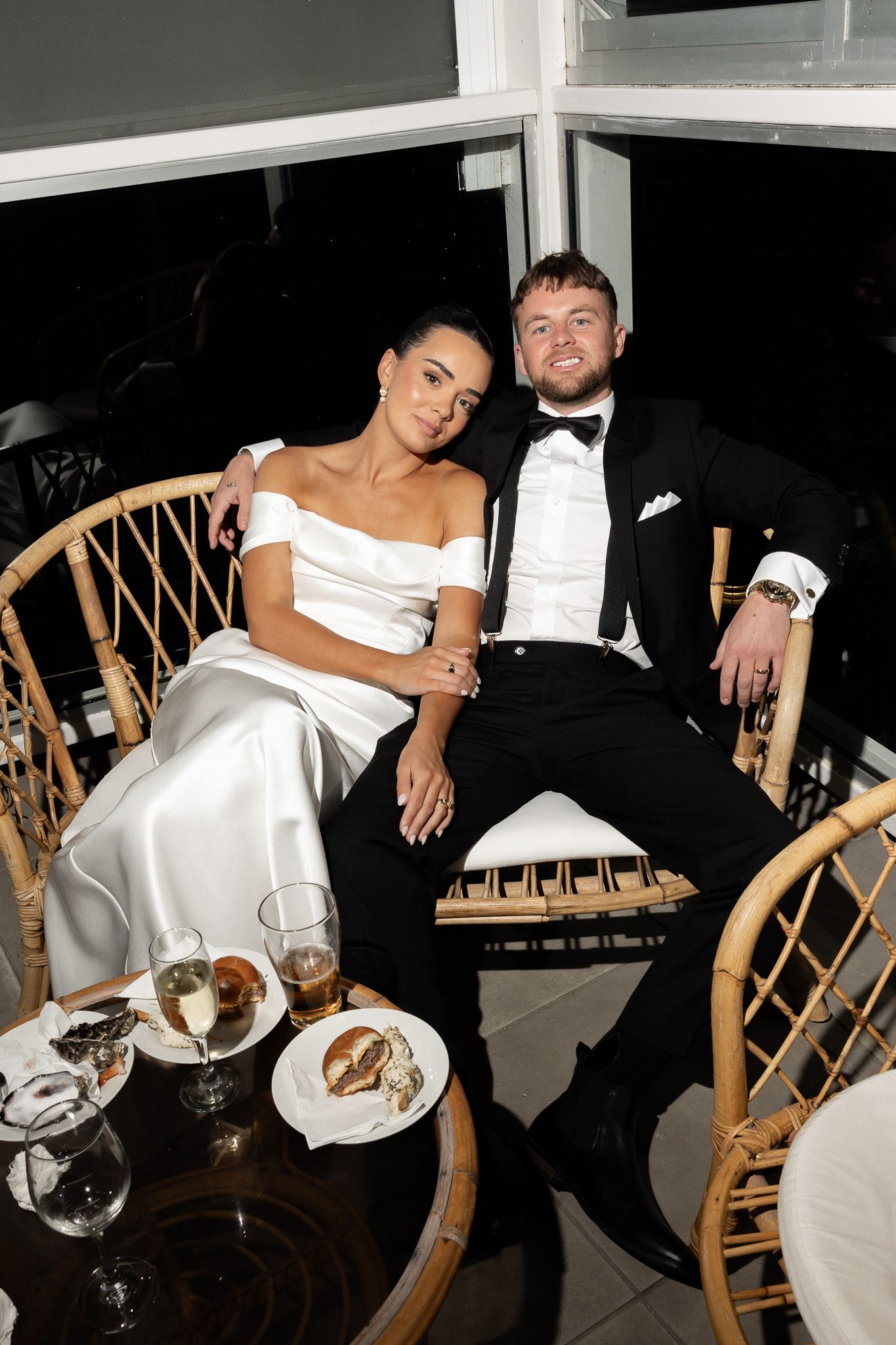 Bride and groom sitting together at wedding reception table, wearing white gown and black tuxedo.